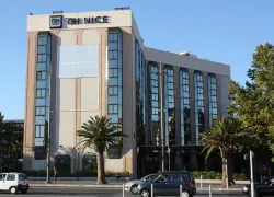 Façade extérieure de l'hôtel NH Nice adaptée pour un séminaire en ville, comprenant une façade beige et vitrée à plusieurs étages avec l'enseigne « NH NICE » au sommet au premier plan, de grands palmiers de part et d'autre de l'entrée au centre, et des voitures et un piéton traversant la rue sous un ciel bleu dégagé en arrière-plan.