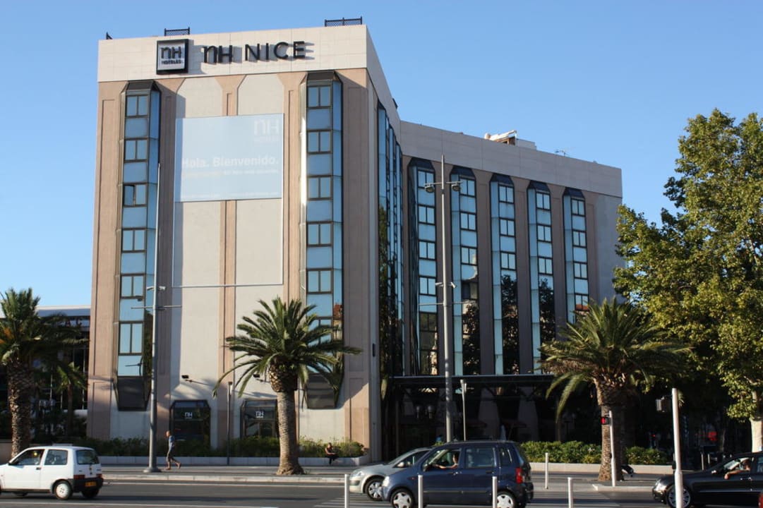 NH Nice hotel exterior fitting for a seminar in the city, featuring beige and glass multi-storey facade with the "NH NICE" signage at the top in the foreground, tall palm trees on either side of the entrance in the centre, and cars and a pedestrian crossing the street under a clear blue sky in the background.