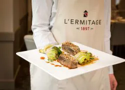 Gros plan de restauration sur place montrant un cuisinier tenant une assiette carrée blanche garnie de tranches de thon poêlé croûté aux graines de sésame et de rouleaux de chou farci, agrémentées d'herbes fraîches et d'un trait de sauce rouge au centre, portant un tablier beige imprimé du logo « L'Ermitage 1897 ».