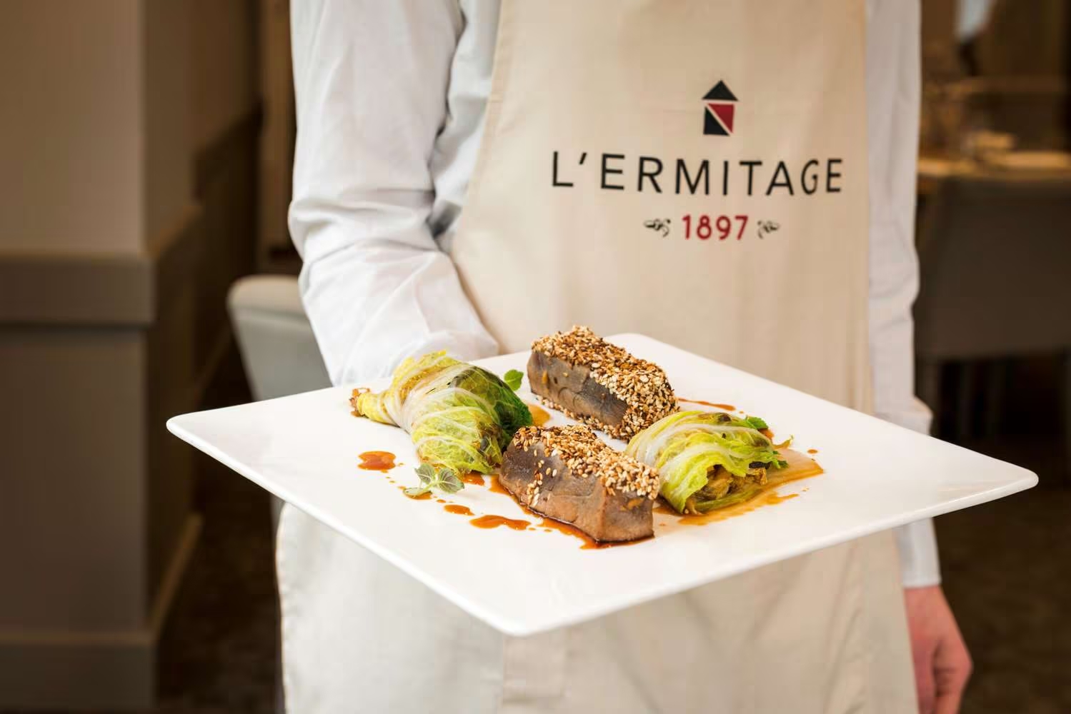 Close-up shot of on-site catering with a chef holding a square white plate of sesame-crusted seared tuna slices alongside stuffed cabbage leaf rolls, garnished with fresh herbs and a red sauce drizzle in the centre, wearing a beige apron printed with the "L'Ermitage 1897" logo.