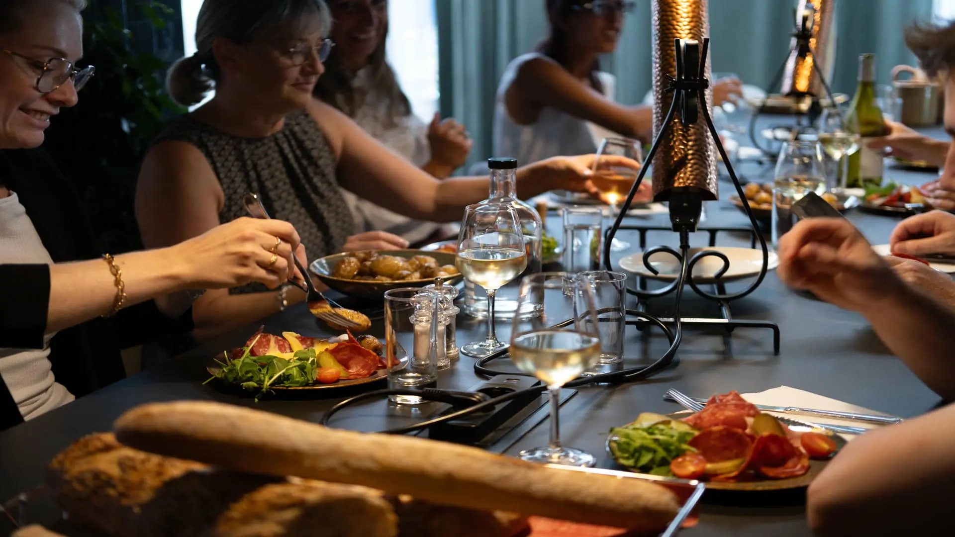en::Restaurant table with traditional raclette grill, bread, cold cuts and potatoes, with guests sharing the seminar meal at Novotel Chartres. fr::Table de restaurant avec appareil à raclette traditionnel, pain, charcuteries et pommes de terre, autour de laquelle des convives partagent un repas séminaire au Novotel Chartres.