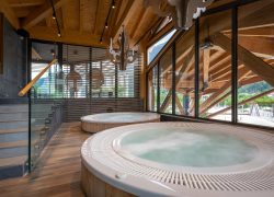 Espace spa d'hôtel avec deux jacuzzis ronds habillés de bois à l'eau fumante au premier plan, lustre décoratif façon bois de cerf suspendu au-dessus, poutres de toit en bois apparentes et escalier avec garde-corps vitré à gauche, et baies vitrées du sol au plafond dévoilant une charpente apparente et une vue sur une ville de montagne à droite, un espace spa adapté à un séminaire bien-être.