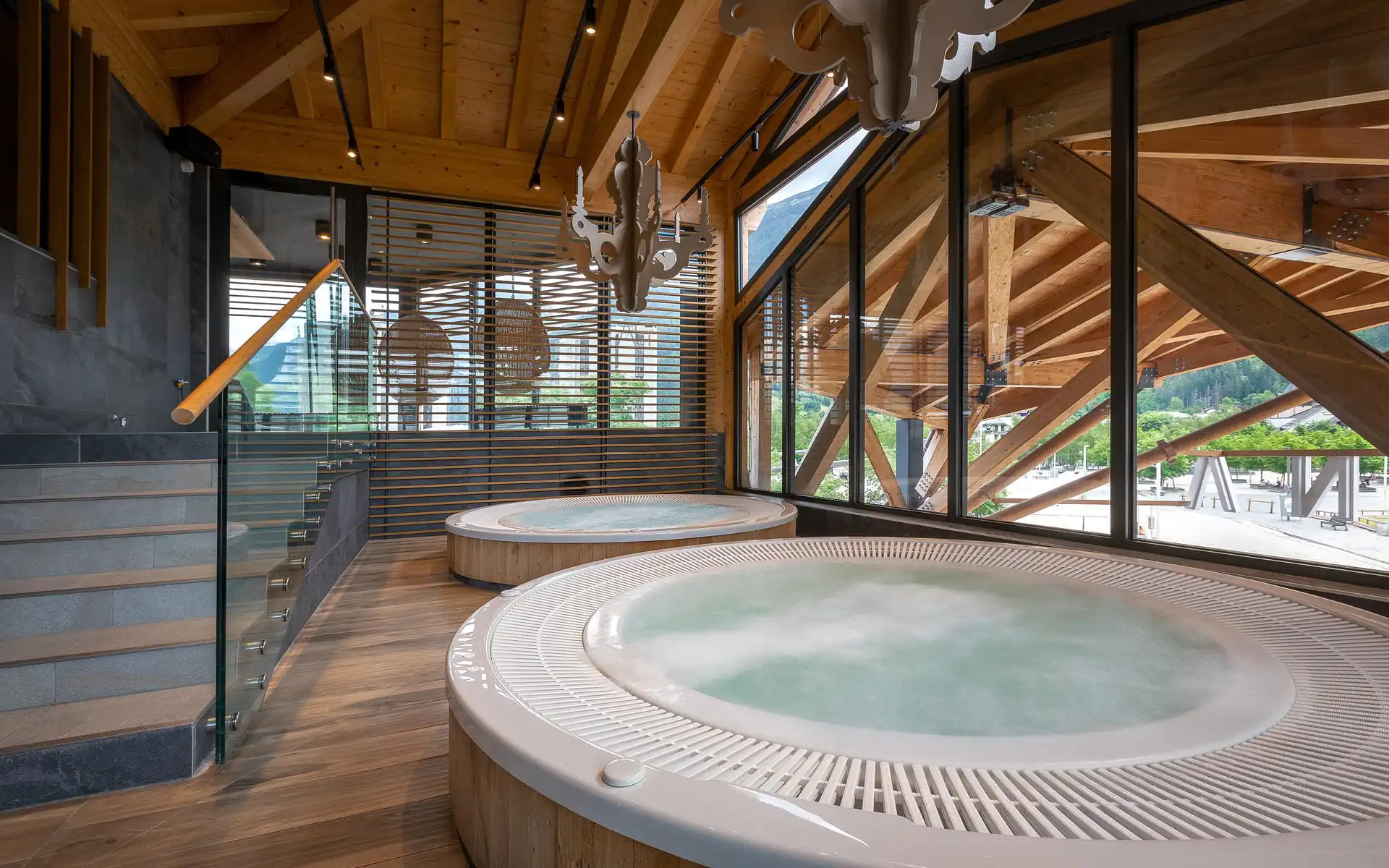 Espace spa d'hôtel avec deux jacuzzis ronds habillés de bois à l'eau fumante au premier plan, lustre décoratif façon bois de cerf suspendu au-dessus, poutres de toit en bois apparentes et escalier avec garde-corps vitré à gauche, et baies vitrées du sol au plafond dévoilant une charpente apparente et une vue sur une ville de montagne à droite, un espace spa adapté à un séminaire bien-être.