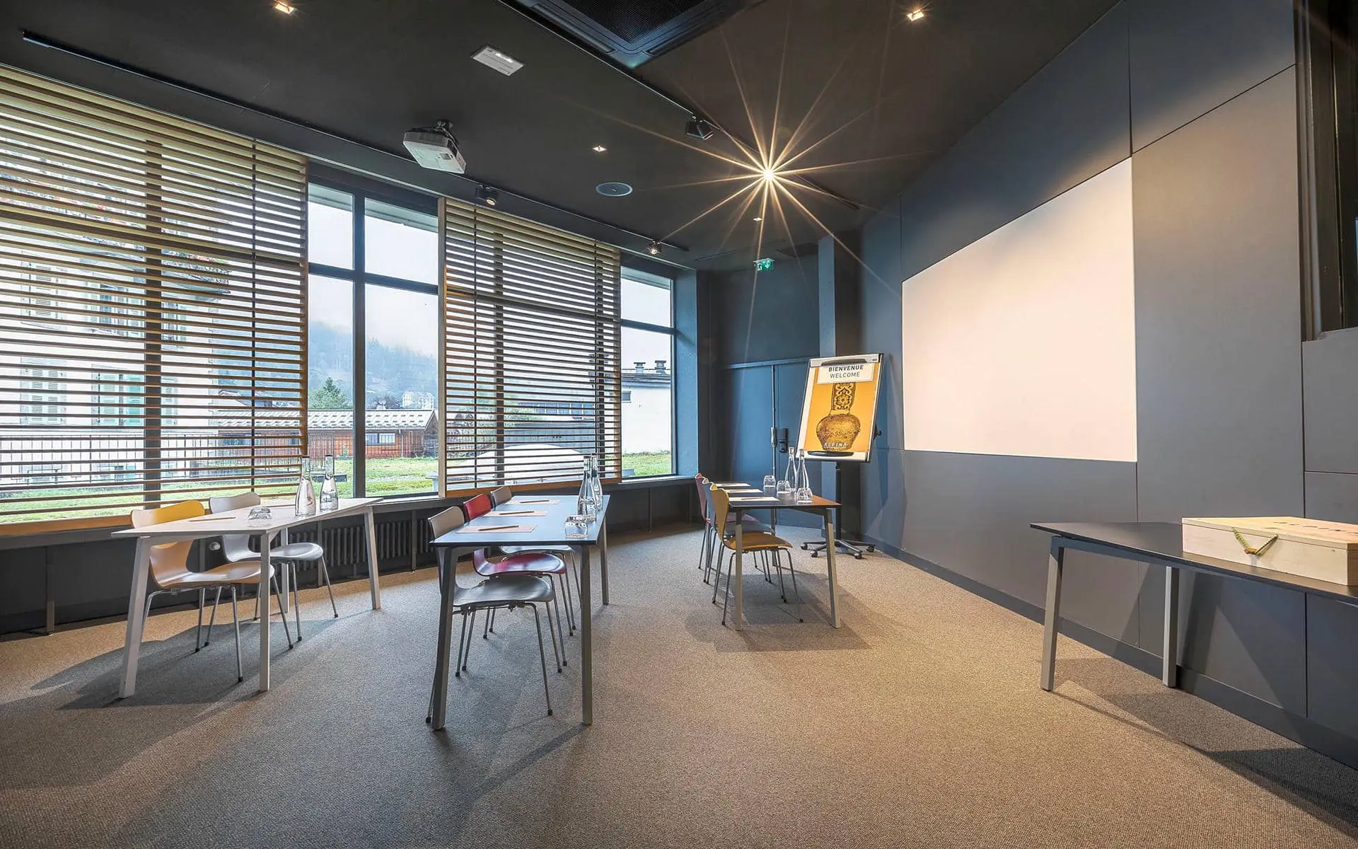 Salle de formation d'hôtel avec des tables individuelles disposées pour une salle de formation, comprenant des blocs-notes, bouteilles en verre et verres sur les tables au premier plan, des chaises colorées au centre, un écran de projection et un paperboard à droite, et de grandes fenêtres avec stores en bois le long du mur de gauche.