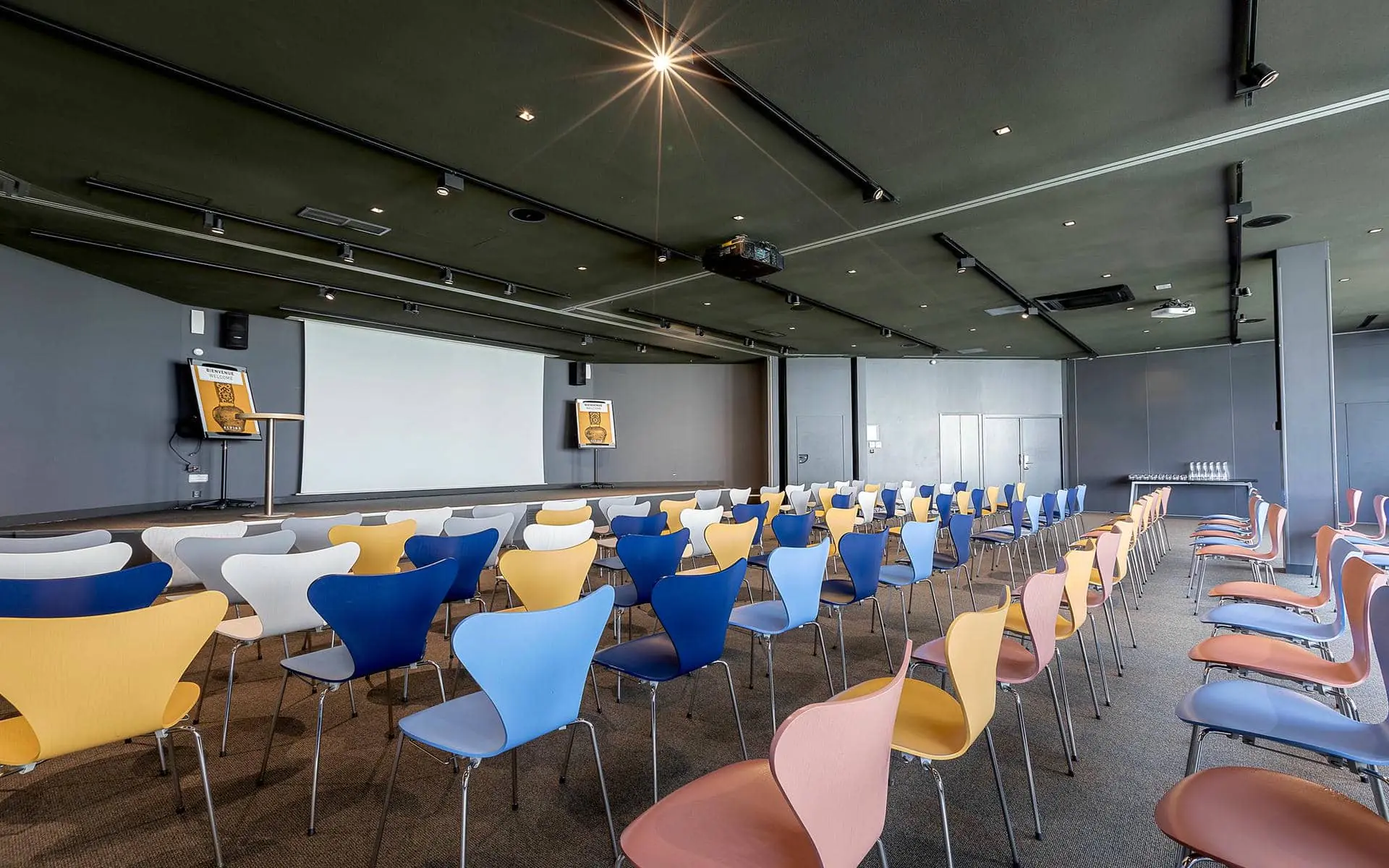 Salle de séminaire d'hôtel avec des rangées de chaises bleues, jaunes, grises et roses au premier plan, une scène avec écran de projection et pupitre sur la gauche, une table de boissons au fond à droite, et un plafond noir avec vidéoprojecteurs suspendus au-dessus pour un hôtel pour séminaire.