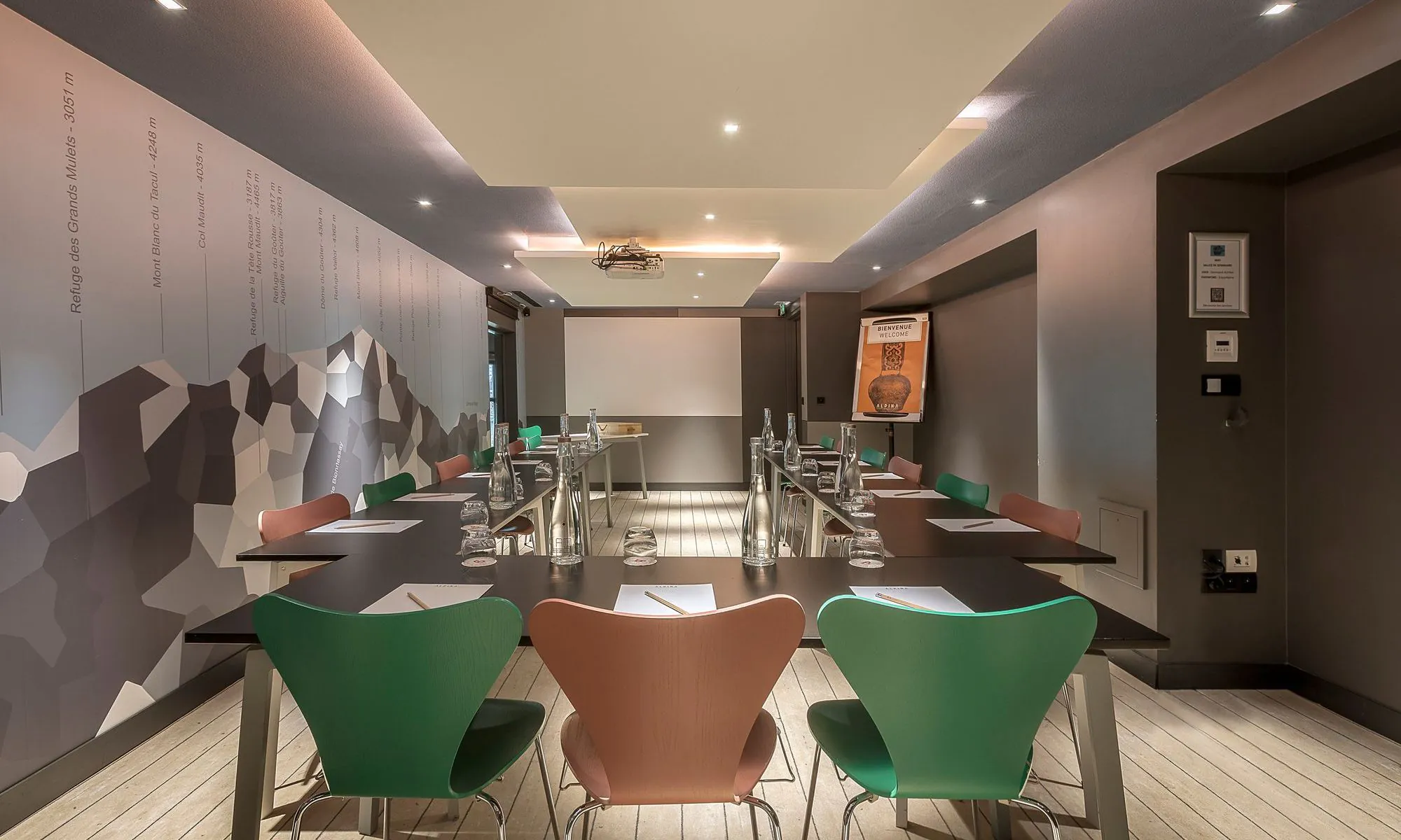Salle de formation d'hôtel avec des tables noires disposées en U pour une salle de formation, comprenant des blocs-notes, crayons, bouteilles en verre et verres au premier plan, des chaises vertes et en bois autour des tables, un écran de projection à l'arrière-plan, et une illustration de montagne couvrant le mur de gauche.