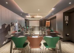 Salle de formation d'hôtel avec des tables noires disposées en U pour une salle de formation, comprenant des blocs-notes, crayons, bouteilles en verre et verres au premier plan, des chaises vertes et en bois autour des tables, un écran de projection à l'arrière-plan, et une illustration de montagne couvrant le mur de gauche.