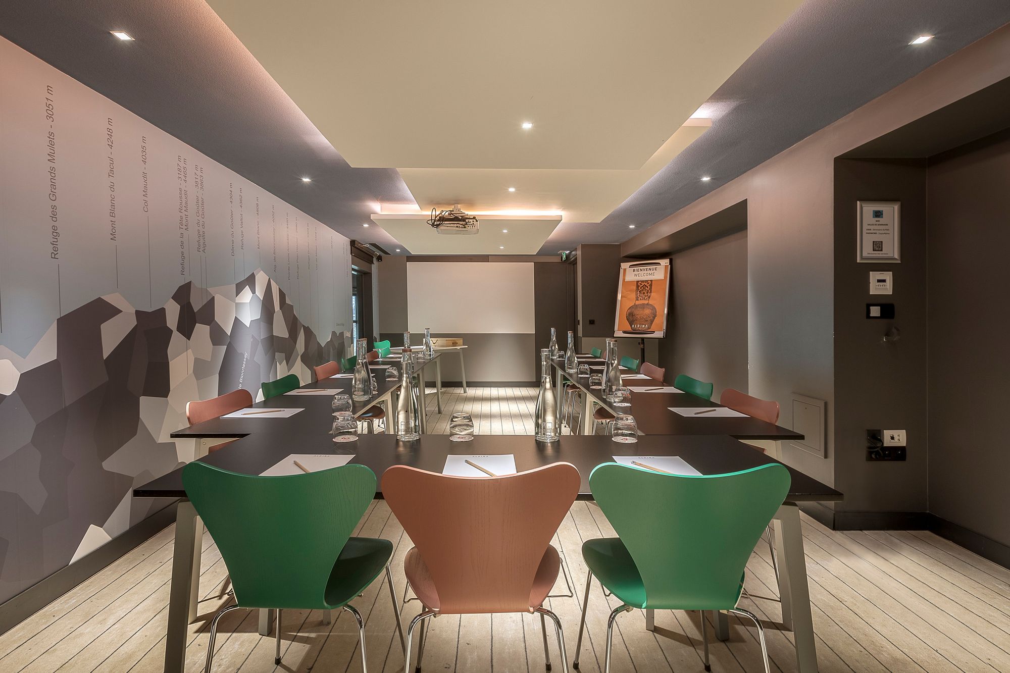 Salle de formation d'hôtel avec des tables noires disposées en U pour une salle de formation, comprenant des blocs-notes, crayons, bouteilles en verre et verres au premier plan, des chaises vertes et en bois autour des tables, un écran de projection à l'arrière-plan, et une illustration de montagne couvrant le mur de gauche.
