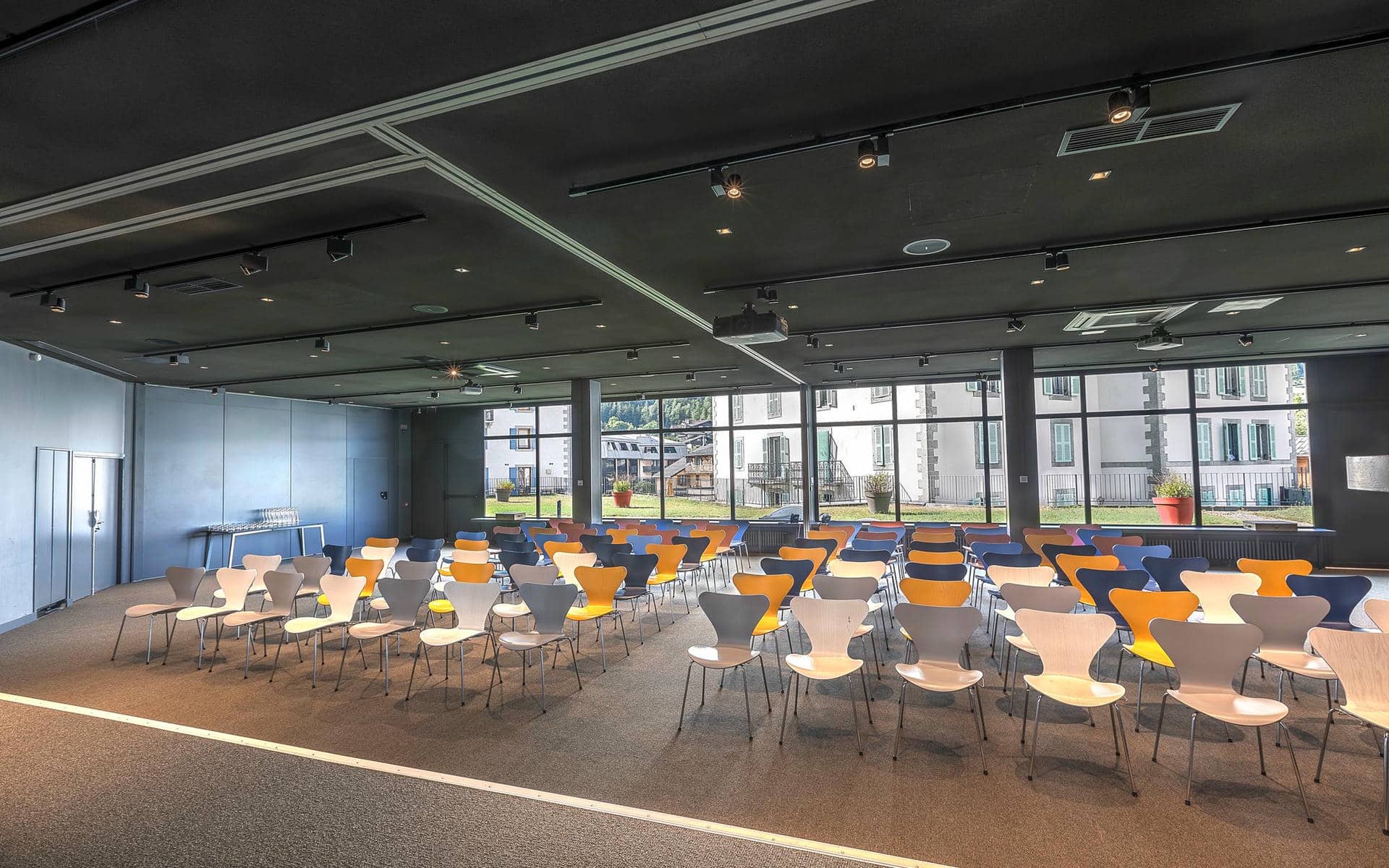 Salle de réunion d'hôtel pour location de salle, avec des rangées de chaises bleues, orange, grises et blanches au premier plan, un vaste espace de présentation ouvert sur la gauche, de grandes baies vitrées sur la droite donnant sur l'extérieur, et un plafond noir avec vidéoprojecteurs suspendus au-dessus.