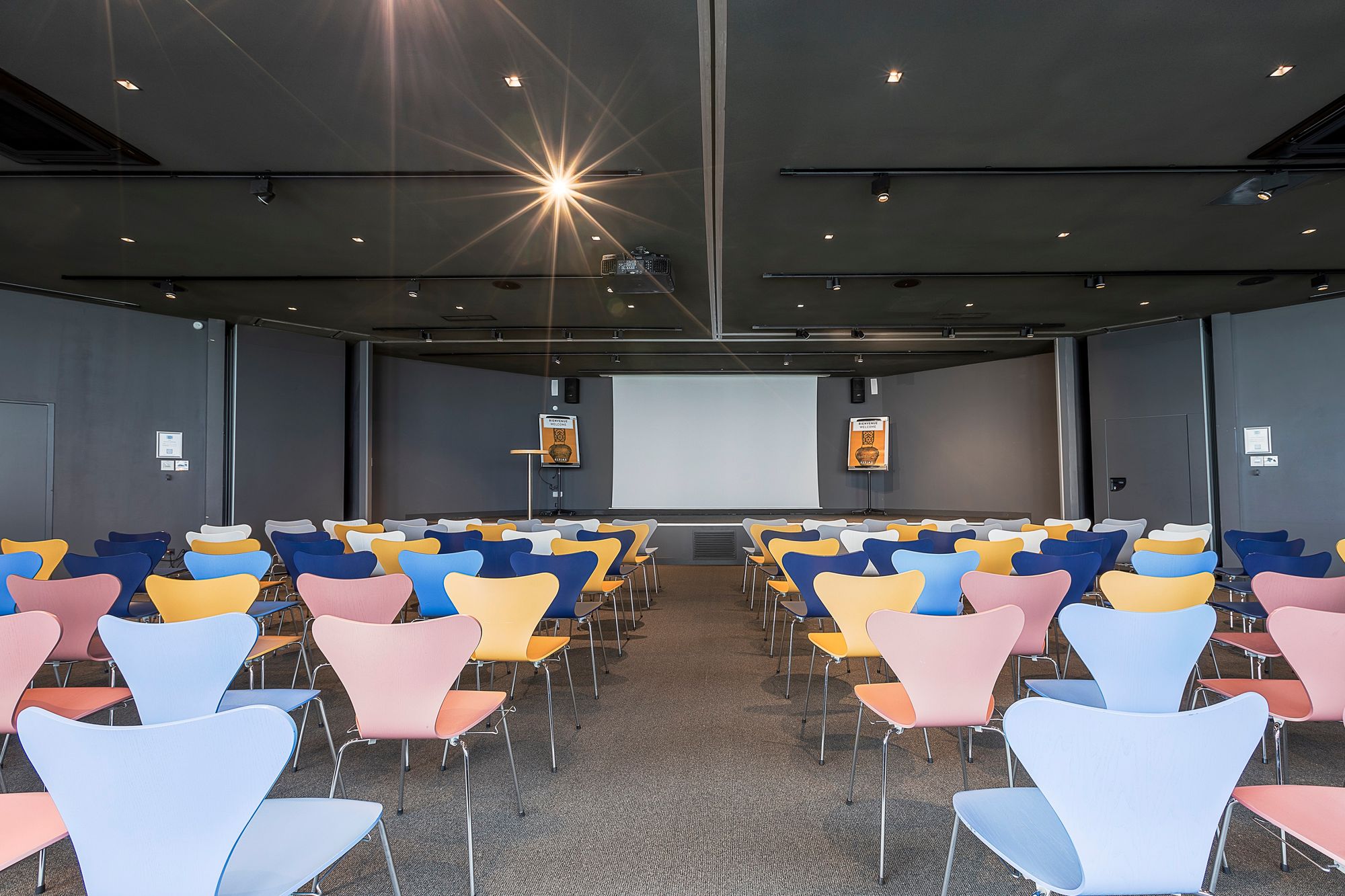 Salle de formation d'hôtel avec des rangées de chaises bleues, jaunes et roses au premier plan, une allée centrale menant à un écran de projection et un pupitre au centre, des murs et un plafond sombres avec éclairage encastré, pour une salle de formation.