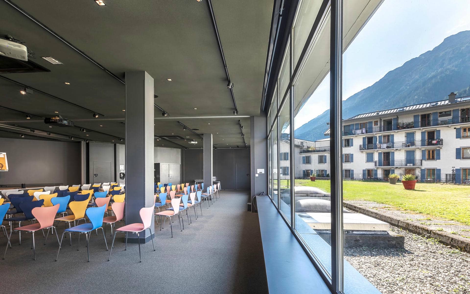 Salle de séminaire pour un séminaire à la montagne, avec des rangées de chaises colorées en plastique moulé rose, bleu, jaune et gris sur une moquette gris foncé au premier plan à gauche, des piliers gris le long d'un couloir avec un écran de projection visible au fond, et des baies vitrées du sol au plafond à droite donnant sur une pelouse, une allée de gravier et des bâtiments de village de montagne aux volets bleus sous une chaîne de montagnes.