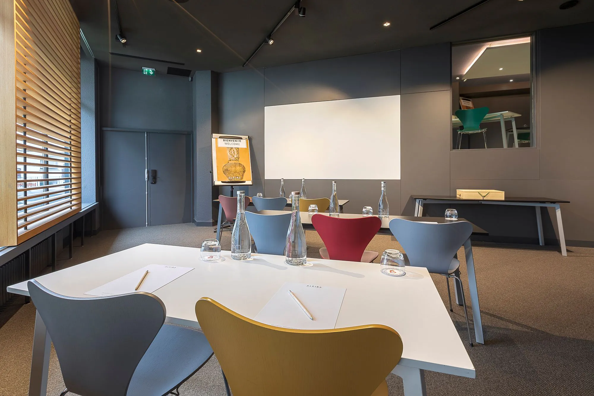 Salle de formation d'hôtel avec des tables blanches disposées en configuration salle de classe pour une salle de formation, comprenant des blocs-notes, crayons, bouteilles en verre et verres au premier plan, des chaises colorées orientées vers un écran de projection au centre, un paperboard à gauche, et de grandes fenêtres avec stores en bois le long du mur de gauche.