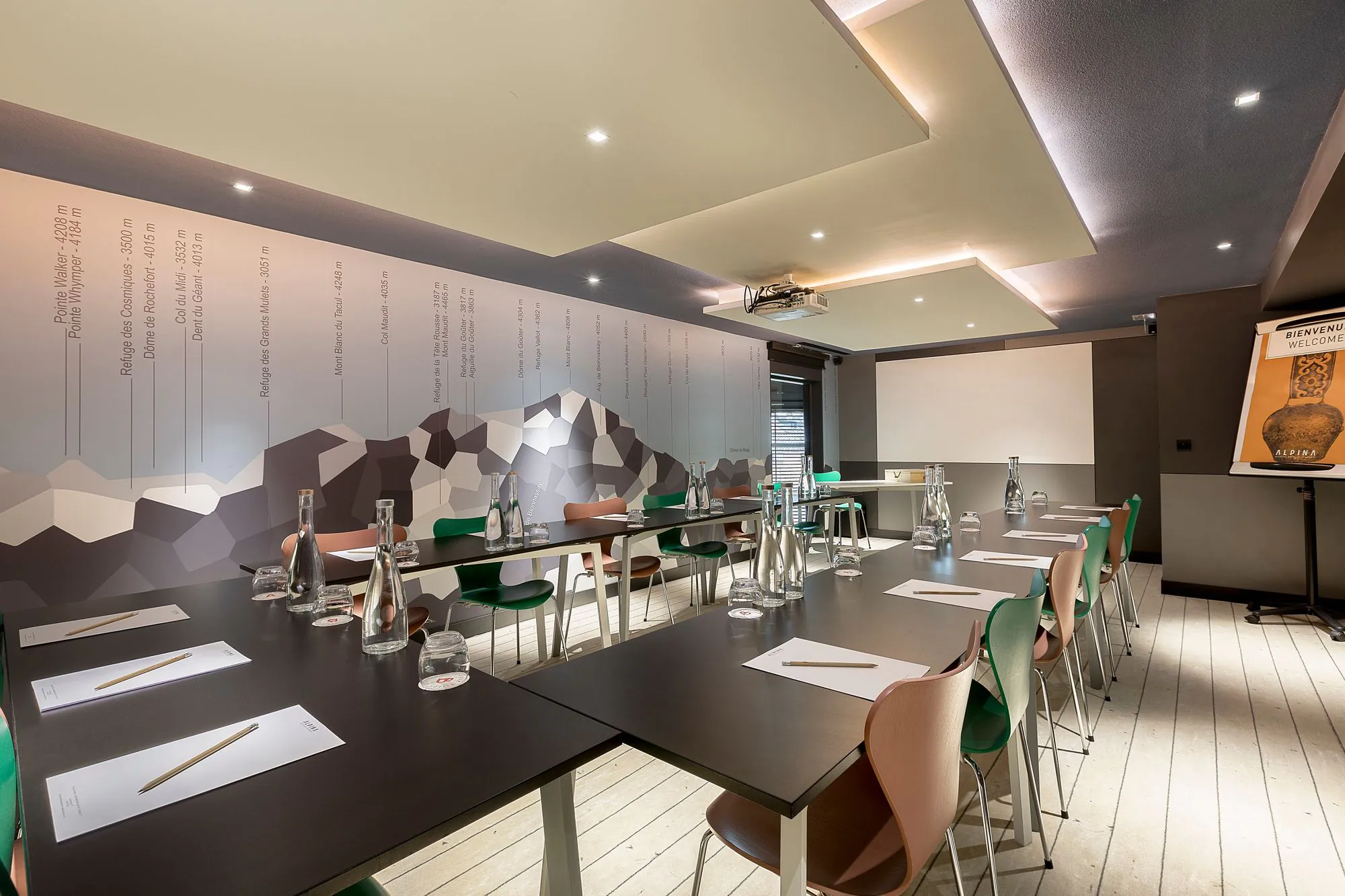 Salle de réunion d'hôtel pour location de salle, avec des tables noires en U équipées de blocs-notes, crayons, bouteilles en verre et verres au premier plan, des chaises vertes et en bois autour des tables, un écran de projection au fond, et une illustration de montagne couvrant le mur de gauche.