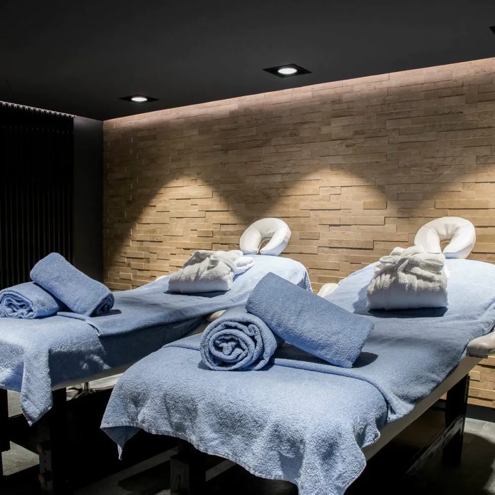 Salle de spa d'hôtel pour séminaire bien-être, avec trois tables de massage recouvertes de serviettes bleu clair au premier plan, des peignoirs pliés sur les tables au centre et au fond, un mur en parement de pierre à l'arrière-plan, un plafond sombre avec éclairage encastré, et une desserte avec produits de soin à gauche.