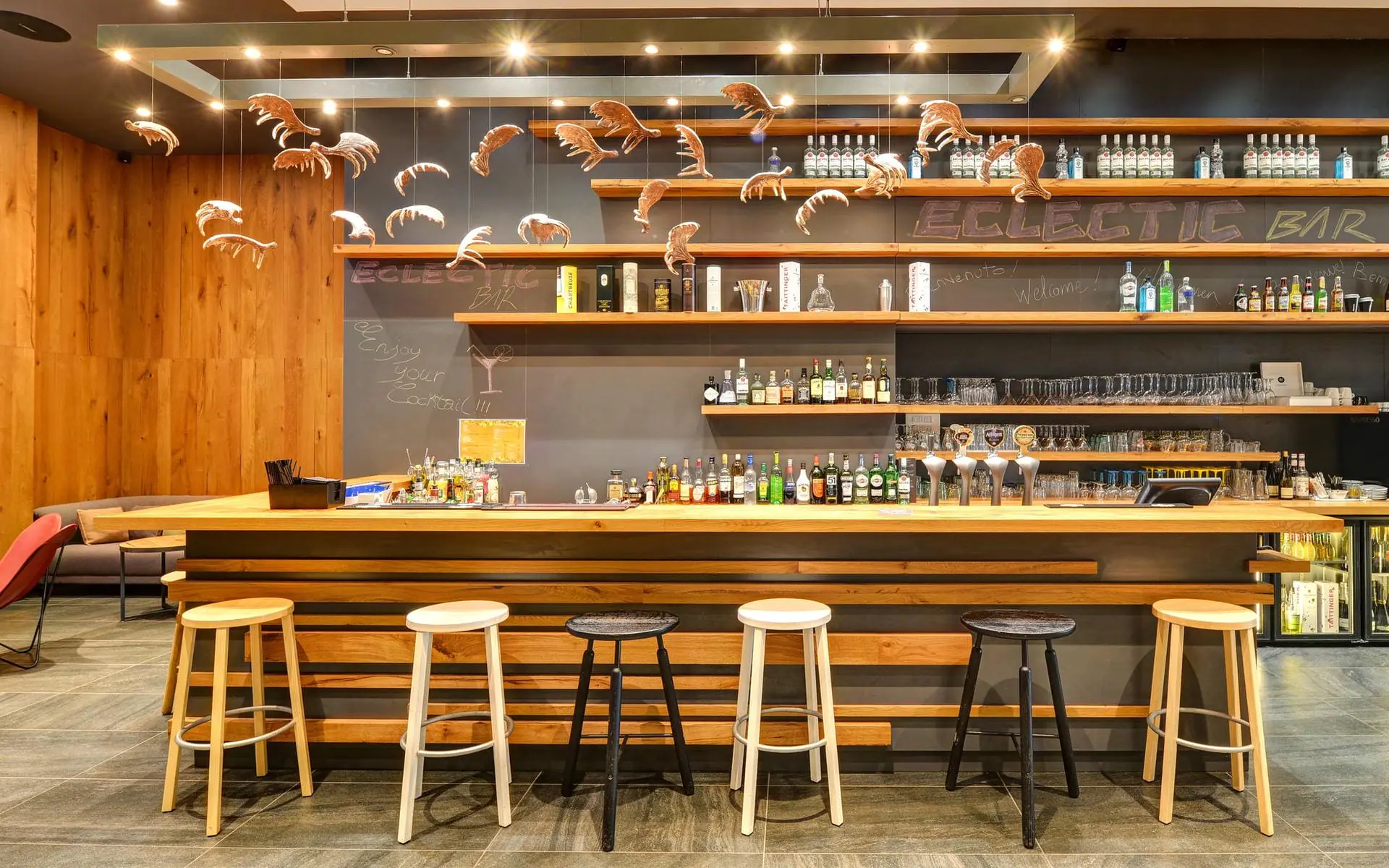 Bar d'hôtel avec comptoir en bois et tabourets hauts dépareillés au premier plan, étagères en bois garnies de bouteilles et verrerie contre un mur en ardoise portant l'inscription « Eclectic Bar » en arrière-plan, et sculptures décoratives en forme de bois de cerf suspendues au plafond au-dessus, un espace bar adapté à un afterwork.