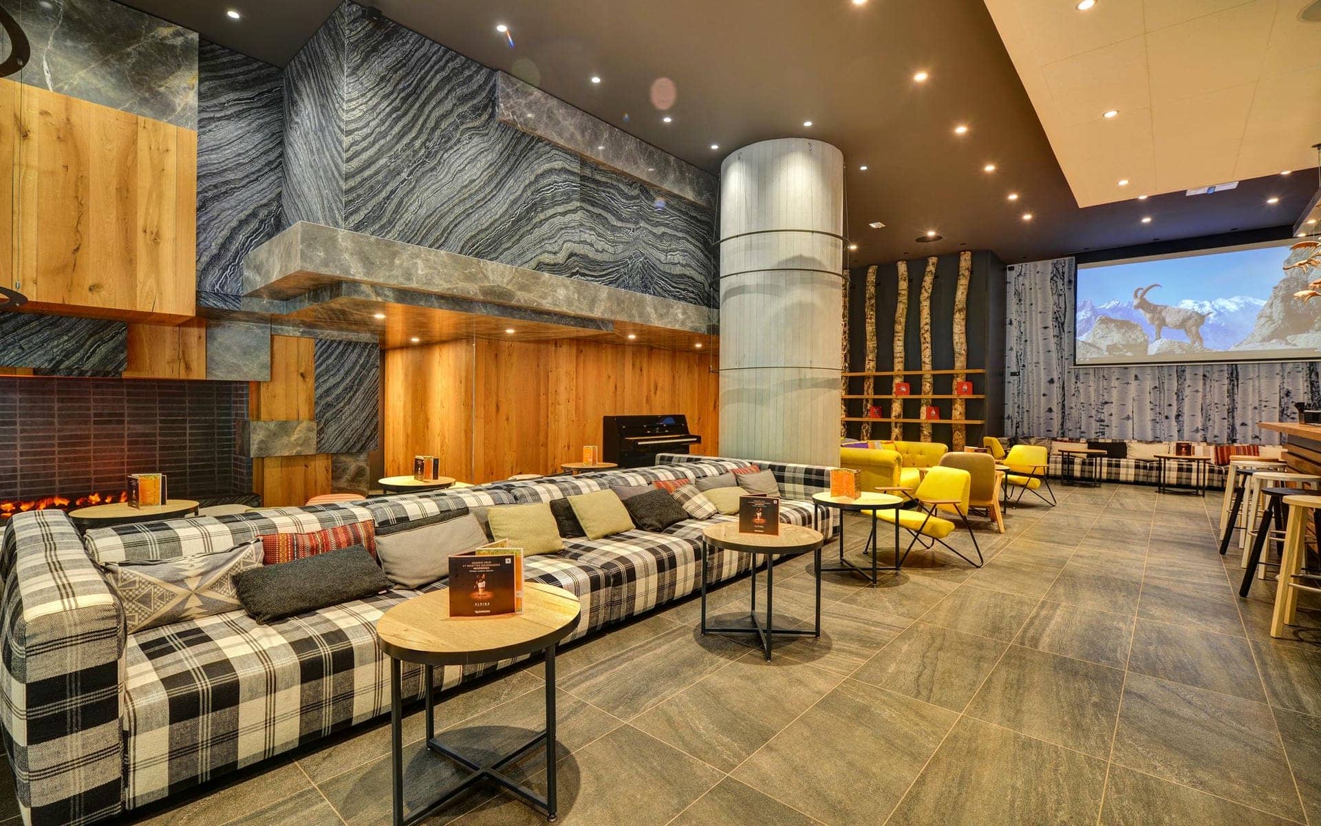 Salon d'hôtel avec long canapé à carreaux et coussins près de tables basses rondes en bois au premier plan, mur habillé de pierre grise et cheminée avec piano droit à gauche, fauteuils jaunes et panneaux muraux effet bouleau au milieu, et écran de projection montrant une scène de bouquetin en montagne près d'un bar avec tabourets hauts à droite, un salon adapté à un séminaire à la montagne.