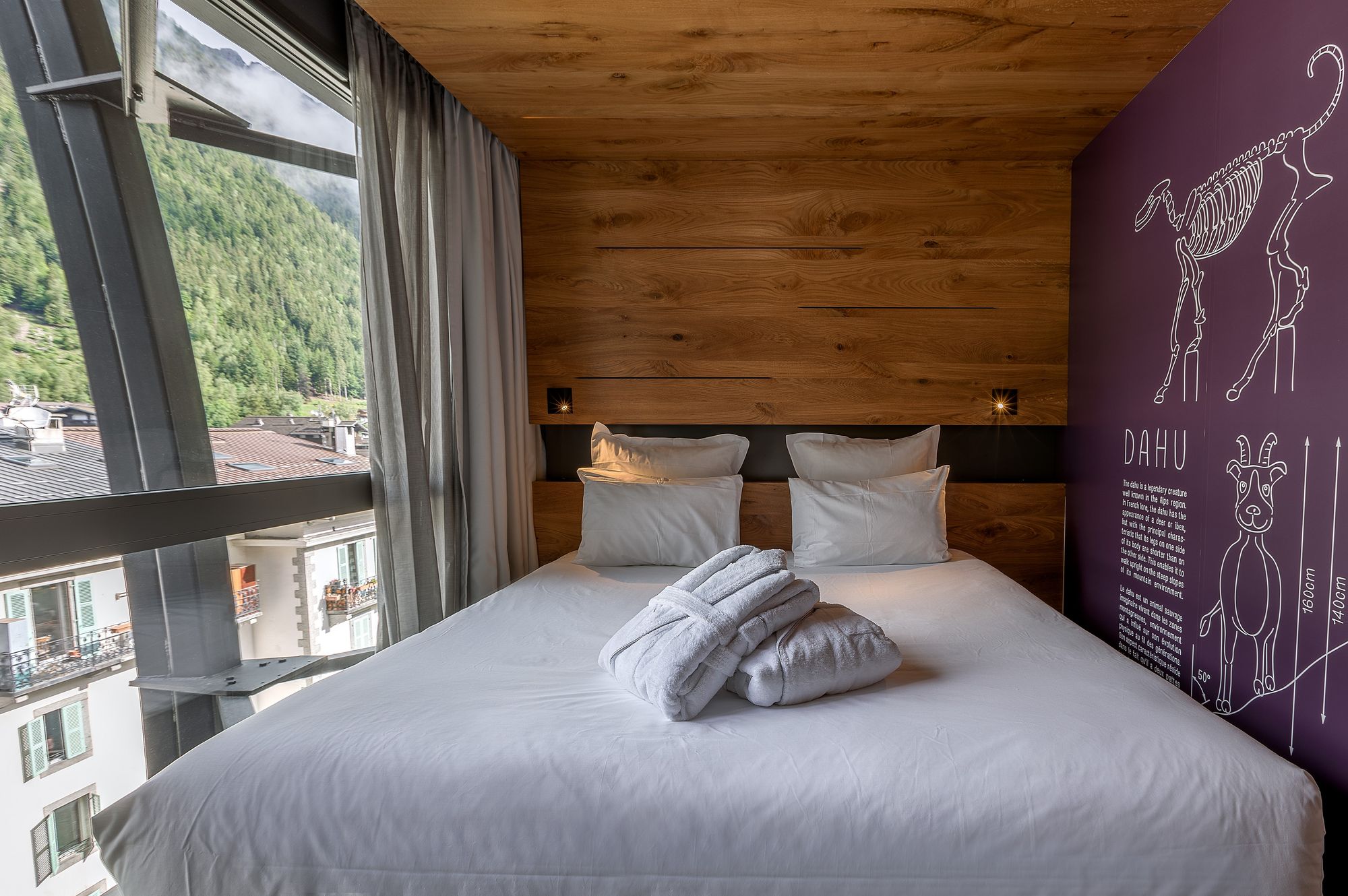 Chambre d'hôtel avec lit double aux draps blancs et tête de lit en panneaux de bois au centre, peignoirs pliés et serviette posés sur le lit au premier plan, mur violet à droite illustré d'un schéma de la créature « Dahu », et grande fenêtre à gauche donnant sur des montagnes boisées et des toits, une chambre adaptée à un séminaire à la montagne.