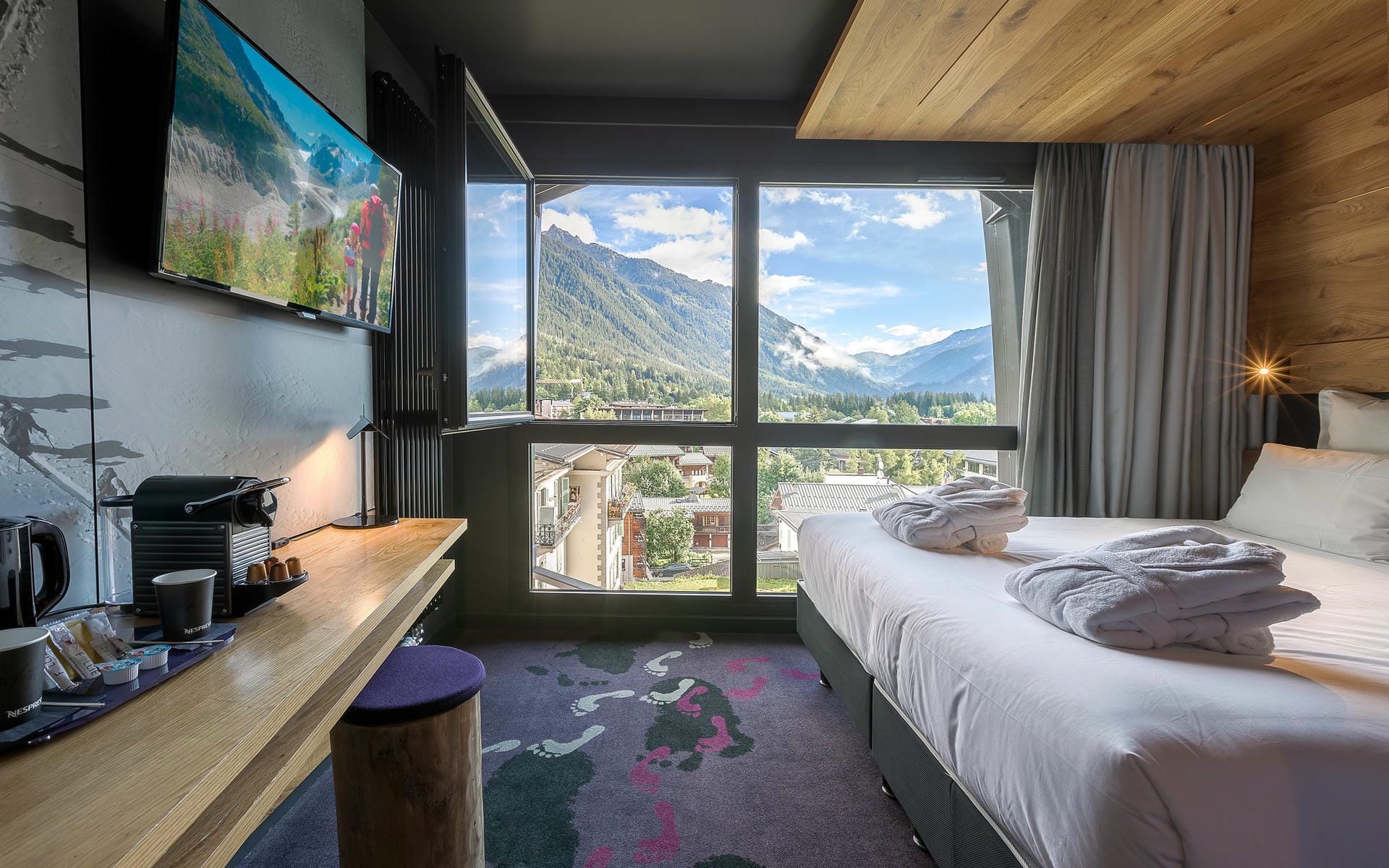 Chambre d'hôtel avec lit aux draps blancs et serviettes et peignoirs pliés à droite, bureau en bois avec machine à café, tasses et télévision murale à gauche, moquette à motif d'empreintes de pas au milieu, et fenêtre ouverte dévoilant des montagnes boisées et une ville de vallée en arrière-plan, une chambre adaptée à un séminaire à la montagne.
