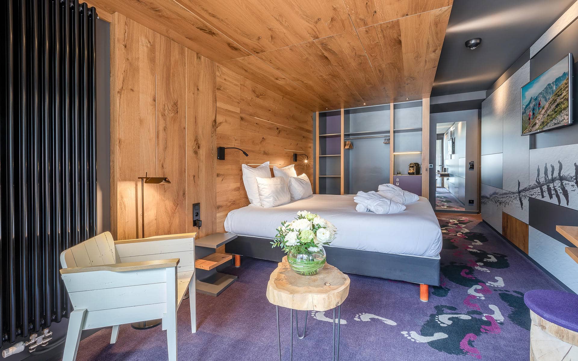 Chambre d'hôtel pour un séminaire à la montagne, avec un lit habillé de linge blanc et des serviettes pliées contre un mur en bois avec une penderie ouverte à droite, une table d'appoint en tranche de tronc portant un bouquet de fleurs blanches au premier plan, un radiateur haut noir et un fauteuil en bois blanc près d'un petit bureau à gauche, et une moquette violette imprimée d'empreintes de pas blanches, roses et noires menant à un couloir avec miroir et télévision murale en arrière-plan.