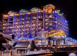 Vue extérieure nocturne de l'hôtel « Alpina » avec sa façade en gradins de style chalet illuminée en bleu et en tons chauds au-dessus d'un porche d'entrée en bois couvert portant le nom de l'hôtel illuminé au premier plan, guirlandes lumineuses soulignant les toits et avant-toits, et passerelle en bois traversant une rivière devant, un hôtel de montagne adapté à un séminaire à la montagne.