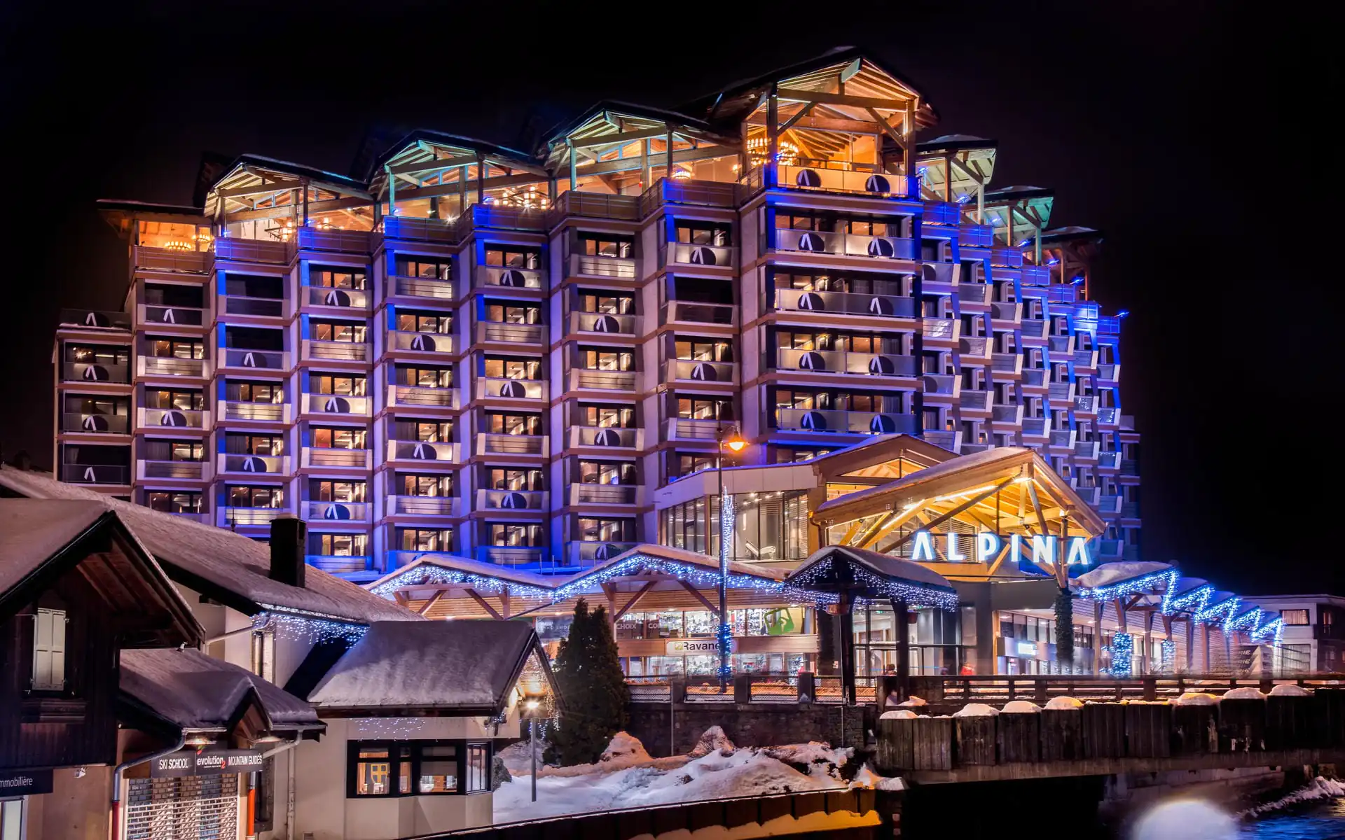Vue extérieure nocturne de l'hôtel « Alpina » avec sa façade en gradins de style chalet illuminée en bleu et en tons chauds au-dessus d'un porche d'entrée en bois couvert portant le nom de l'hôtel illuminé au premier plan, guirlandes lumineuses soulignant les toits et avant-toits, et passerelle en bois traversant une rivière devant, un hôtel de montagne adapté à un séminaire à la montagne.