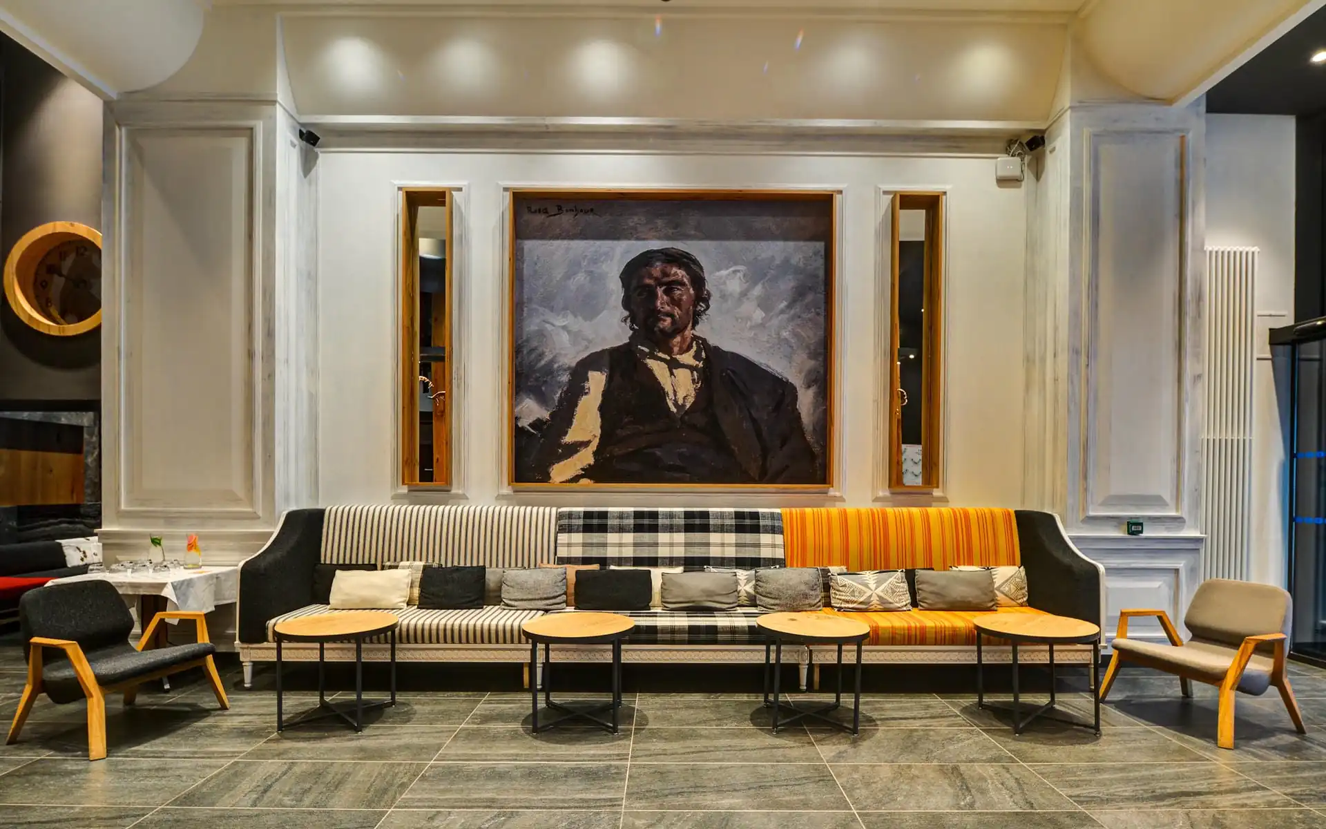Salon d'hôtel avec long canapé aux sections d'assise rayées, à carreaux et orange rayée et coussins à motifs au premier plan, tables basses rondes en bois sur pieds métalliques noirs devant, fauteuils à structure bois de part et d'autre, grand tableau de portrait encadré sur un mur blanc à panneaux au-dessus, et horloge murale ronde en bois à gauche, un salon adapté à un séminaire à la montagne.