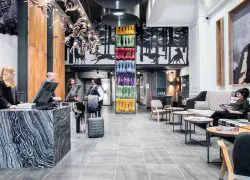 Lobby d'hôtel avec comptoir de réception habillé de marbre gris foncé et personnel assistant les clients au premier plan, colonne enveloppée de cordes multicolores et fresque murale représentant des loups en course au-dessus, sculptures décoratives en forme de bois de cerf suspendues au plafond à gauche, et canapé rayé et à carreaux avec tableau de portrait encadré à droite, un lobby accueillant pour un séminaire à la montagne.
