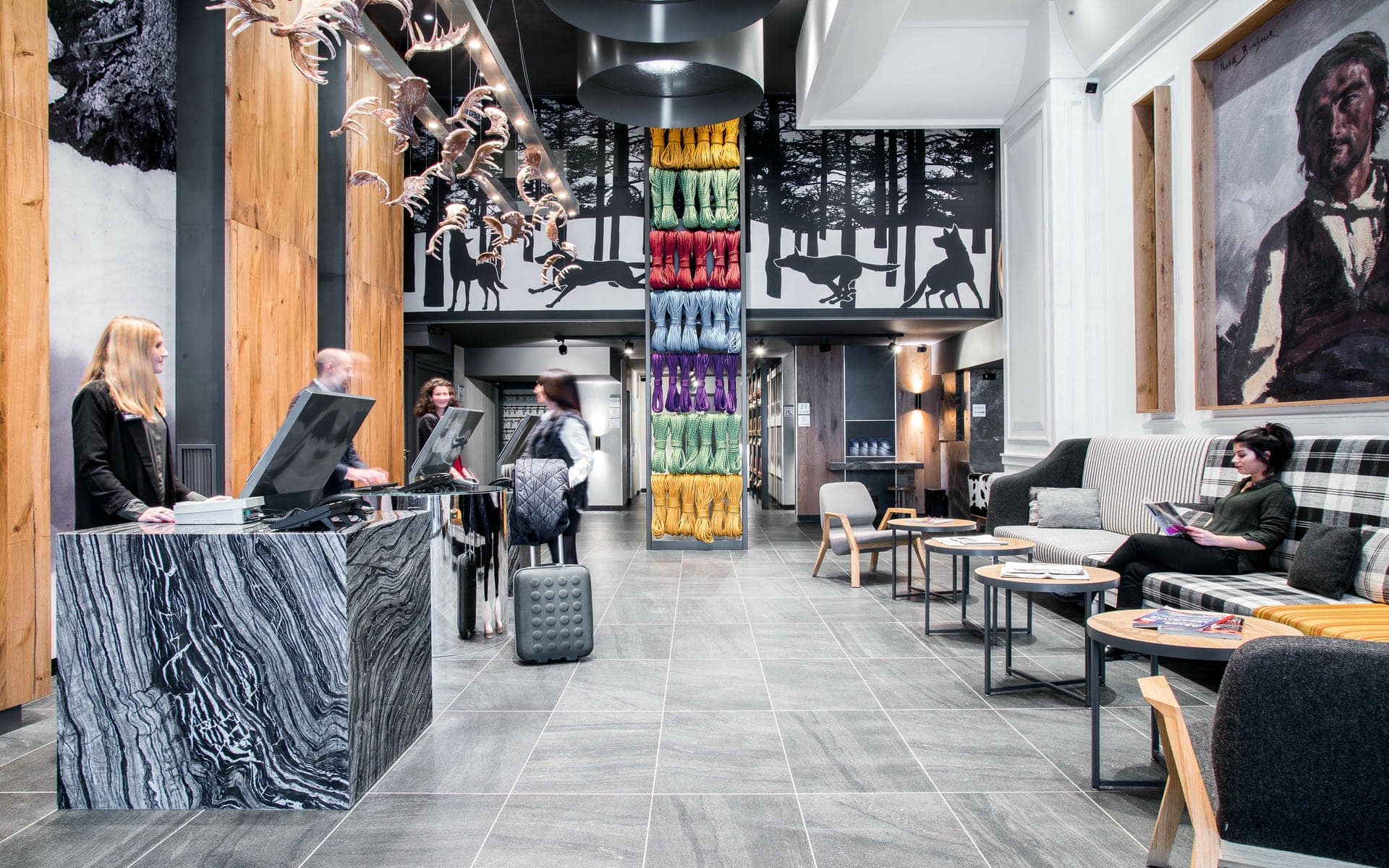 Lobby d'hôtel avec comptoir de réception habillé de marbre gris foncé et personnel assistant les clients au premier plan, colonne enveloppée de cordes multicolores et fresque murale représentant des loups en course au-dessus, sculptures décoratives en forme de bois de cerf suspendues au plafond à gauche, et canapé rayé et à carreaux avec tableau de portrait encadré à droite, un lobby accueillant pour un séminaire à la montagne.