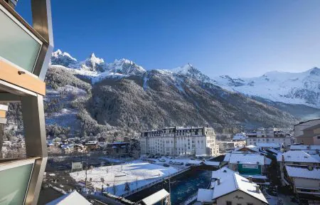 Vue depuis un balcon d'hôtel avec garde-corps en métal et bois à gauche, surplombant la ville enneigée de Chamonix avec un grand bâtiment historique blanc et une rivière traversant le centre, et sommets montagneux enneigés et déchiquetés s'élevant au-dessus de versants boisés sous un ciel bleu dégagé en arrière-plan, une vue panoramique adaptée à un séminaire à la montagne.