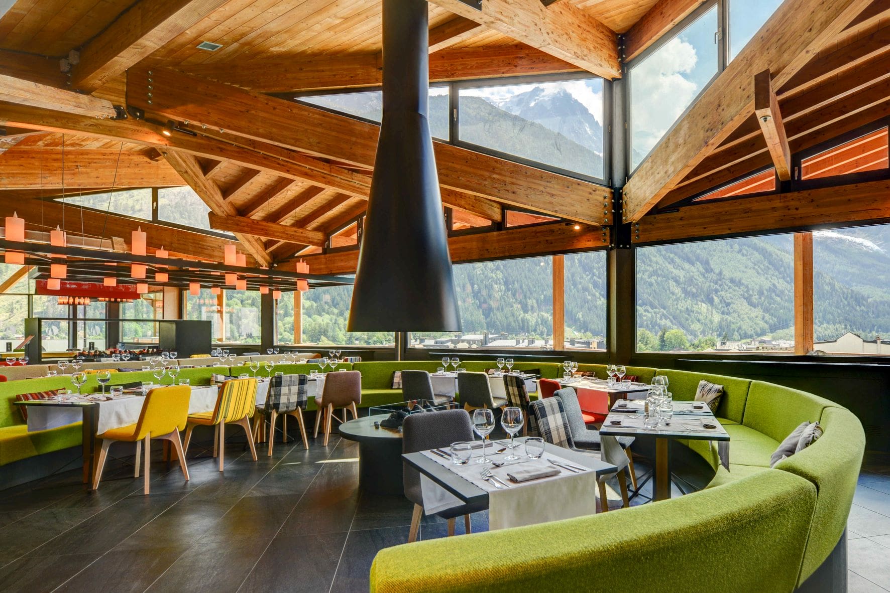 Restaurant d'hôtel avec banquette verte incurvée entourant des tables nappées de blanc dressées de verres à vin et chaises à motifs colorés au premier plan, grande cheminée suspendue conique noire au centre, poutres de toit en bois apparentes et suspensions en forme de lanternes orange au-dessus, et baies vitrées du sol au plafond dévoilant des montagnes boisées à droite, un restaurant proposant une restauration sur place dans un cadre de séminaire à la montagne.