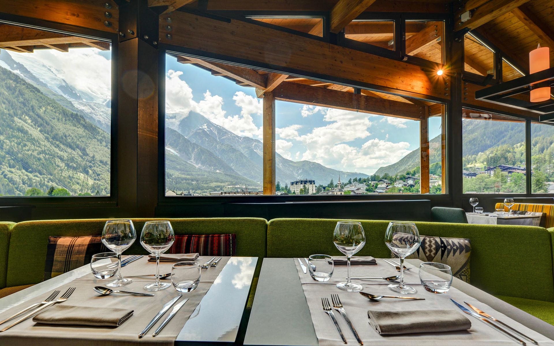 Restaurant d'hôtel avec banquettes rembourrées vertes et tables nappées de gris dressées de verres à vin, couverts et serviettes pliées au premier plan, poutres en bois apparentes encadrant de grandes baies vitrées donnant sur un village de montagne avec clocher d'église et sommets enneigés en arrière-plan, et second espace repas visible à droite, un restaurant proposant une restauration sur place avec une vue de séminaire à la montagne.