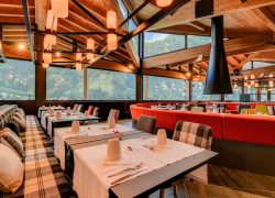 Restaurant d'hôtel avec banquette à carreaux près de tables nappées de blanc dressées de tasses et couverts au premier plan, banquette rouge incurvée avec chaises à motifs colorés au milieu, cheminée suspendue conique noire et suspensions en forme de lanternes accrochées aux poutres de toit en bois au-dessus, et baies vitrées du sol au plafond donnant sur des montagnes boisées en arrière-plan, un restaurant proposant une restauration sur place dans un cadre de séminaire à la montagne.