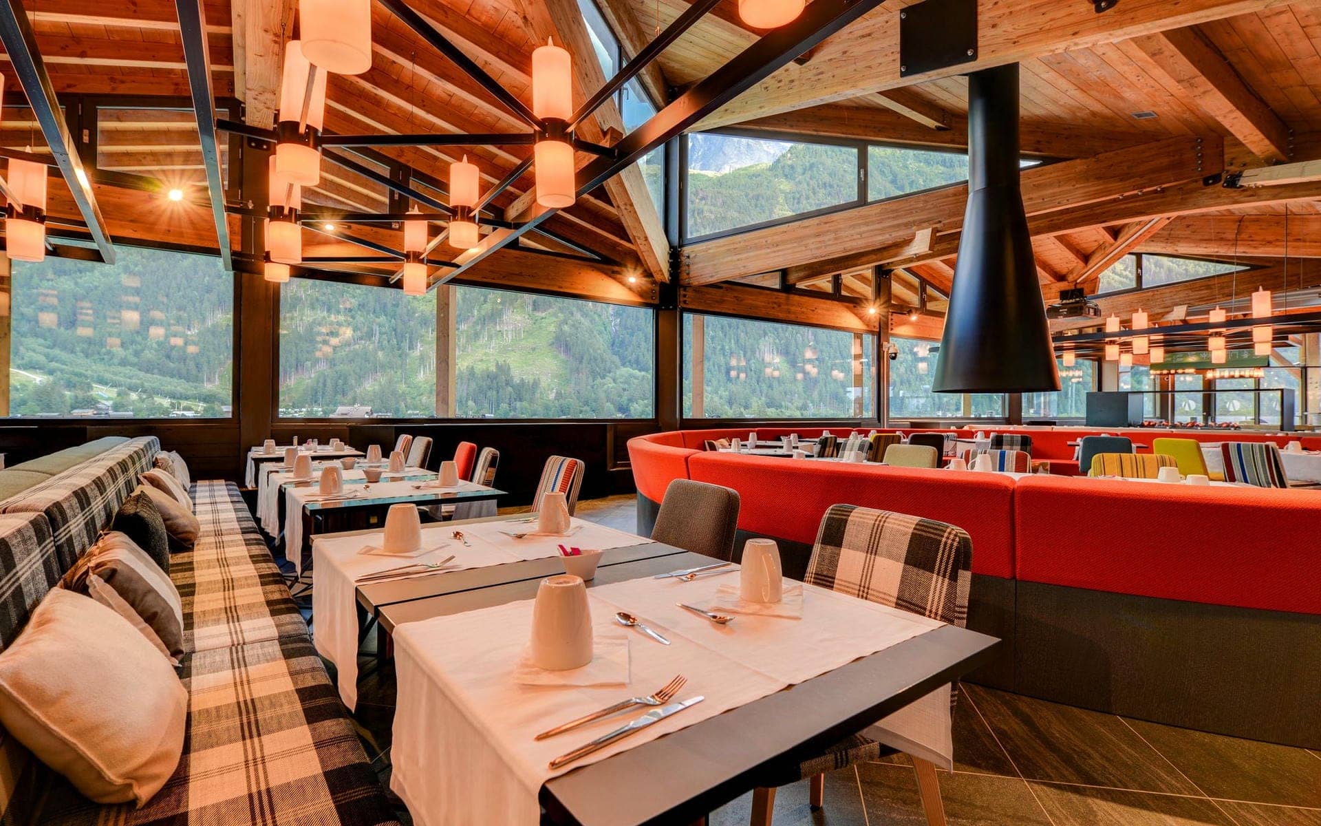 Restaurant d'hôtel avec banquette à carreaux près de tables nappées de blanc dressées de tasses et couverts au premier plan, banquette rouge incurvée avec chaises à motifs colorés au milieu, cheminée suspendue conique noire et suspensions en forme de lanternes accrochées aux poutres de toit en bois au-dessus, et baies vitrées du sol au plafond donnant sur des montagnes boisées en arrière-plan, un restaurant proposant une restauration sur place dans un cadre de séminaire à la montagne.