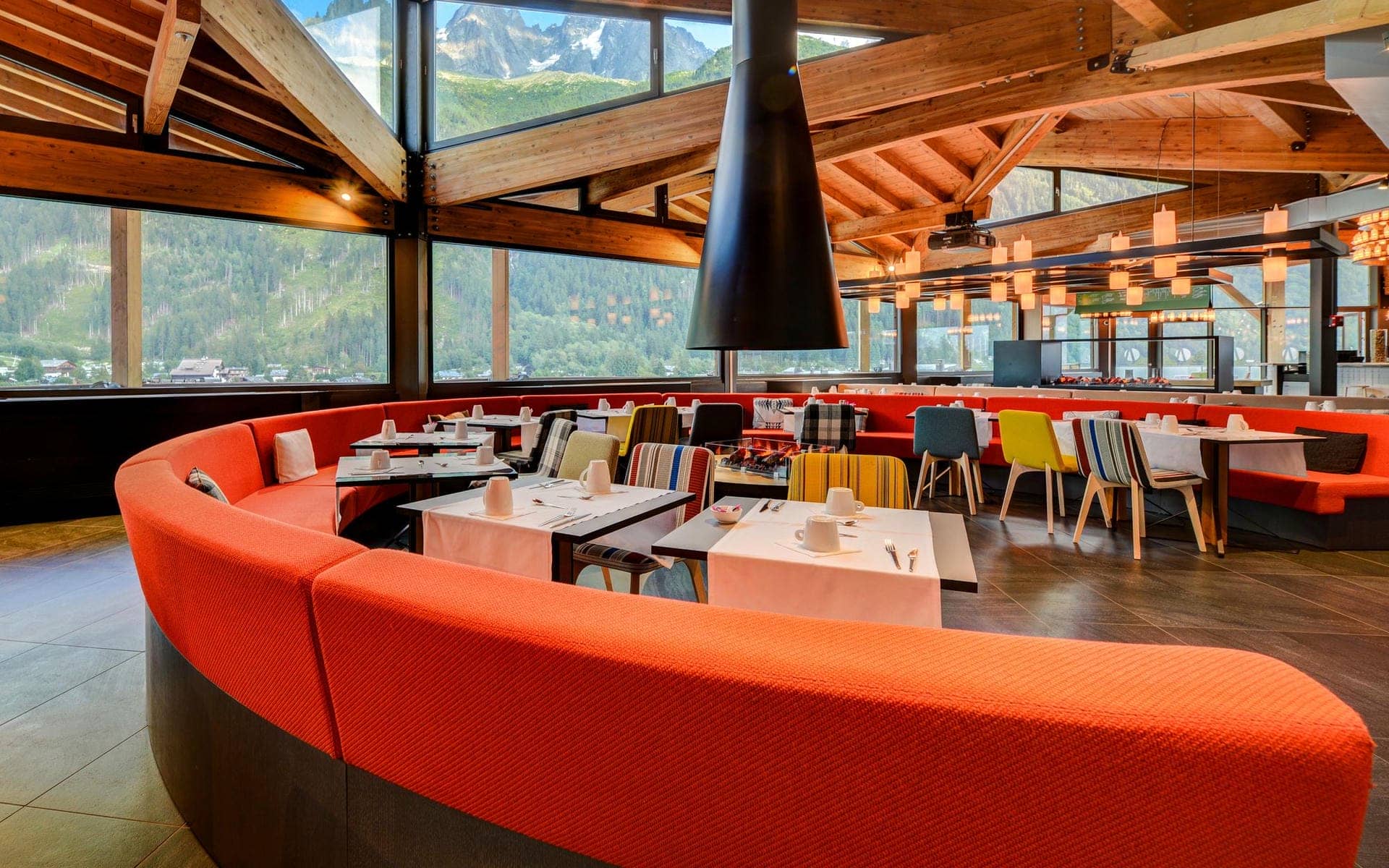 Restaurant d'hôtel avec banquette orange incurvée entourant des tables nappées de blanc dressées de tasses et couverts au premier plan, chaises à motifs colorés autour de tables supplémentaires au milieu, grande cheminée suspendue conique noire et suspensions en forme de lanternes au-dessus, et baies vitrées du sol au plafond dévoilant des montagnes boisées et un sommet enneigé en arrière-plan, un restaurant proposant une restauration sur place dans un cadre de séminaire à la montagne.
