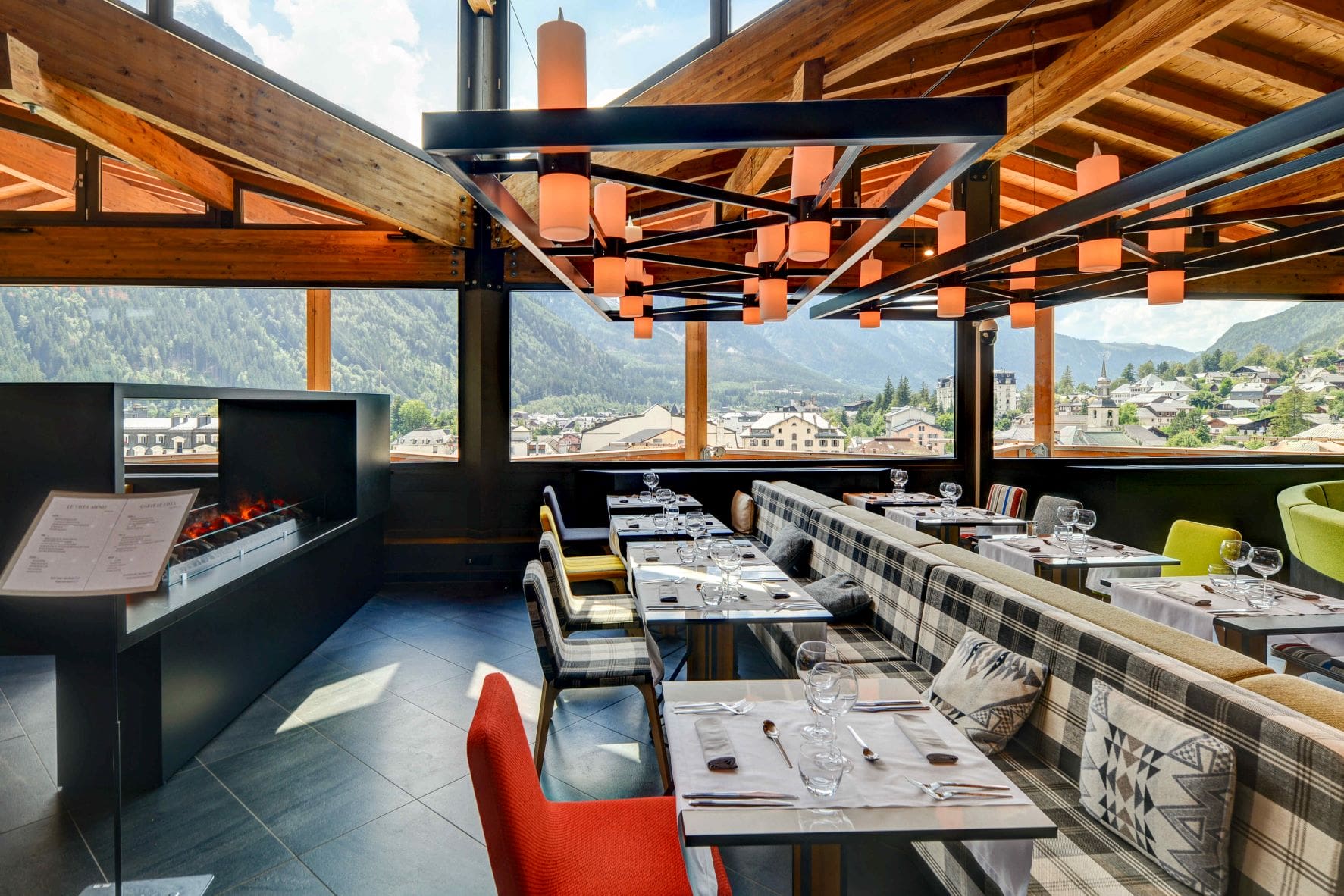 Restaurant d'hôtel avec banquette à carreaux et chaises colorées autour de tables nappées de blanc dressées de verres à vin au premier plan, cheminée noire encastrée et présentoir à menu à gauche, suspension lumineuse en forme de lanternes orange et poutres de toit en bois apparentes au-dessus, et baies vitrées du sol au plafond donnant sur un village de montagne en arrière-plan, un restaurant proposant une restauration sur place avec une vue de séminaire à la montagne.