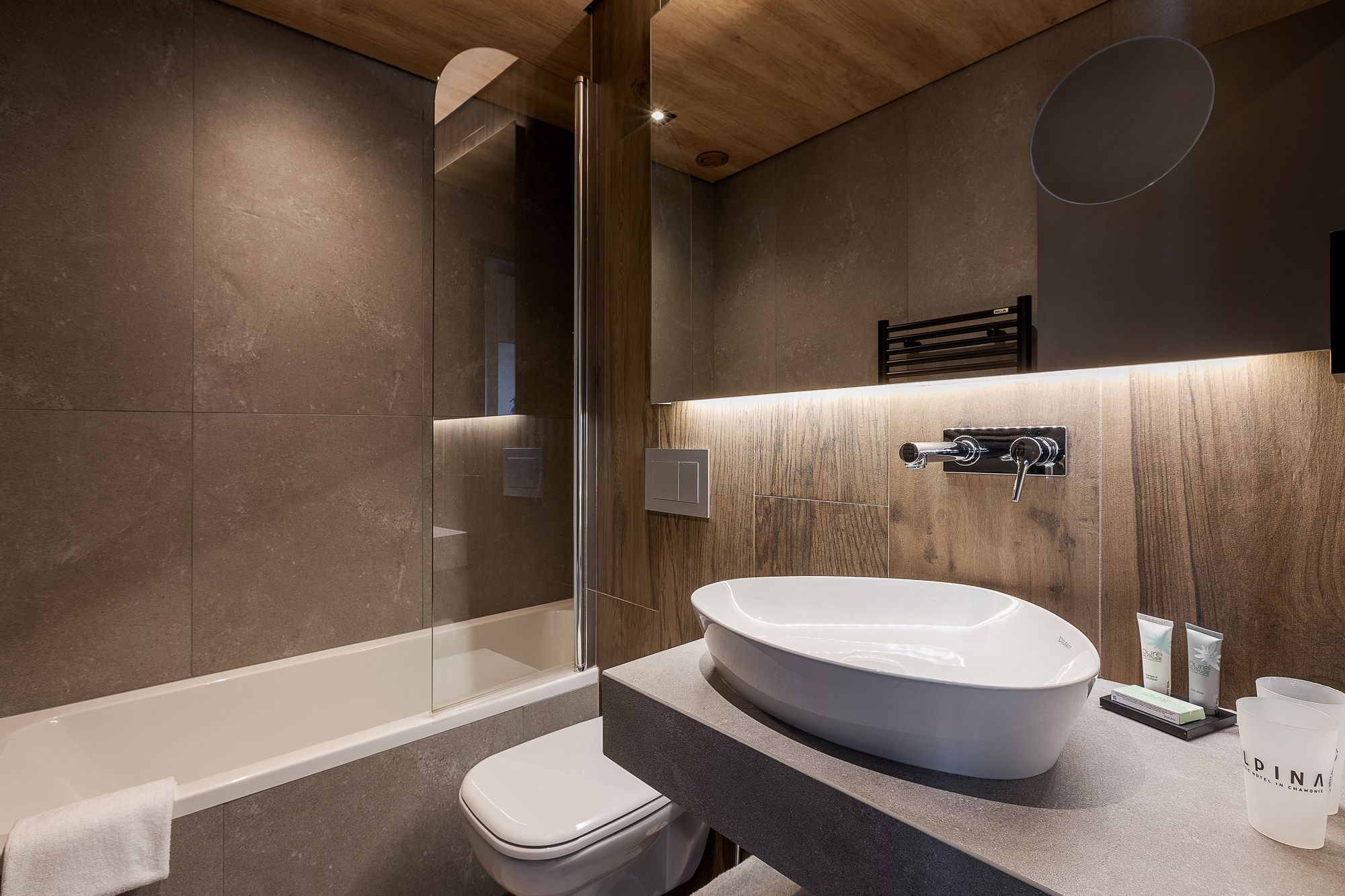 Salle de bain d'hôtel pour un séminaire à la montagne, avec une vasque ovale blanche à poser et un robinet chromé mural sur un plan vasque gris foncé au premier plan, des gobelets siglés et des tubes de produits d'accueil à droite, des toilettes suspendues en dessous, et une baignoire avec paroi vitrée derrière des carreaux effet pierre taupe et un plafond en bois à gauche.