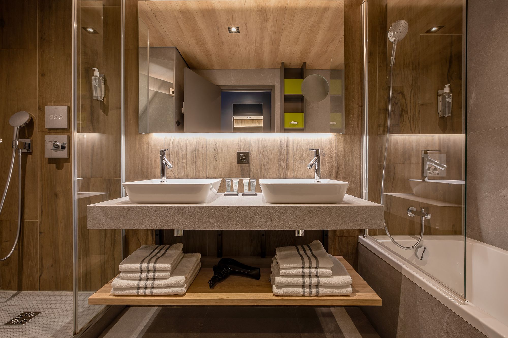 Salle de bain d'hôtel pour un hébergement groupe, avec un double plan vasque doté de deux lavabos blancs à poser et de robinets chromés au premier plan, des serviettes blanches pliées et un sèche-cheveux sur une étagère en bois en dessous, un mur en bois avec un grand miroir rétroéclairé au-dessus, et une douche à l'italienne à gauche et une baignoire avec paroi vitrée à droite.