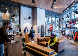 Boutique de matériel de ski avec personne assise sur un canapé en cuir marron essayant des chaussures près d'un banc en bois au premier plan, comptoirs de vente et personnel assistant des clients pour casques et équipement de ski au milieu, étagères présentant vestes, bonnets et accessoires à droite, et suspensions noires accrochées à un plafond en bois au-dessus, une boutique adaptée à un séminaire à la montagne.