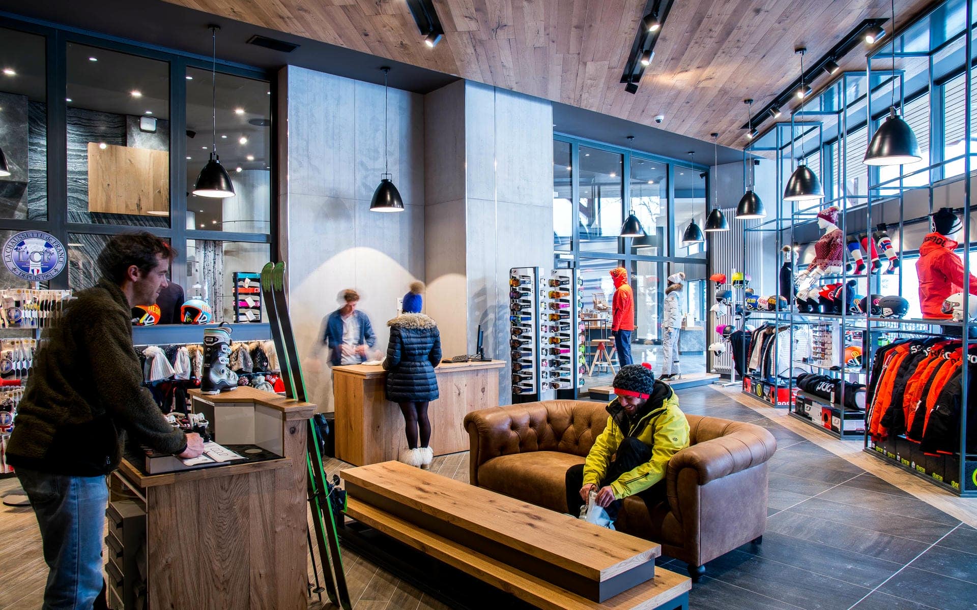 Boutique de matériel de ski avec personne assise sur un canapé en cuir marron essayant des chaussures près d'un banc en bois au premier plan, comptoirs de vente et personnel assistant des clients pour casques et équipement de ski au milieu, étagères présentant vestes, bonnets et accessoires à droite, et suspensions noires accrochées à un plafond en bois au-dessus, une boutique adaptée à un séminaire à la montagne.
