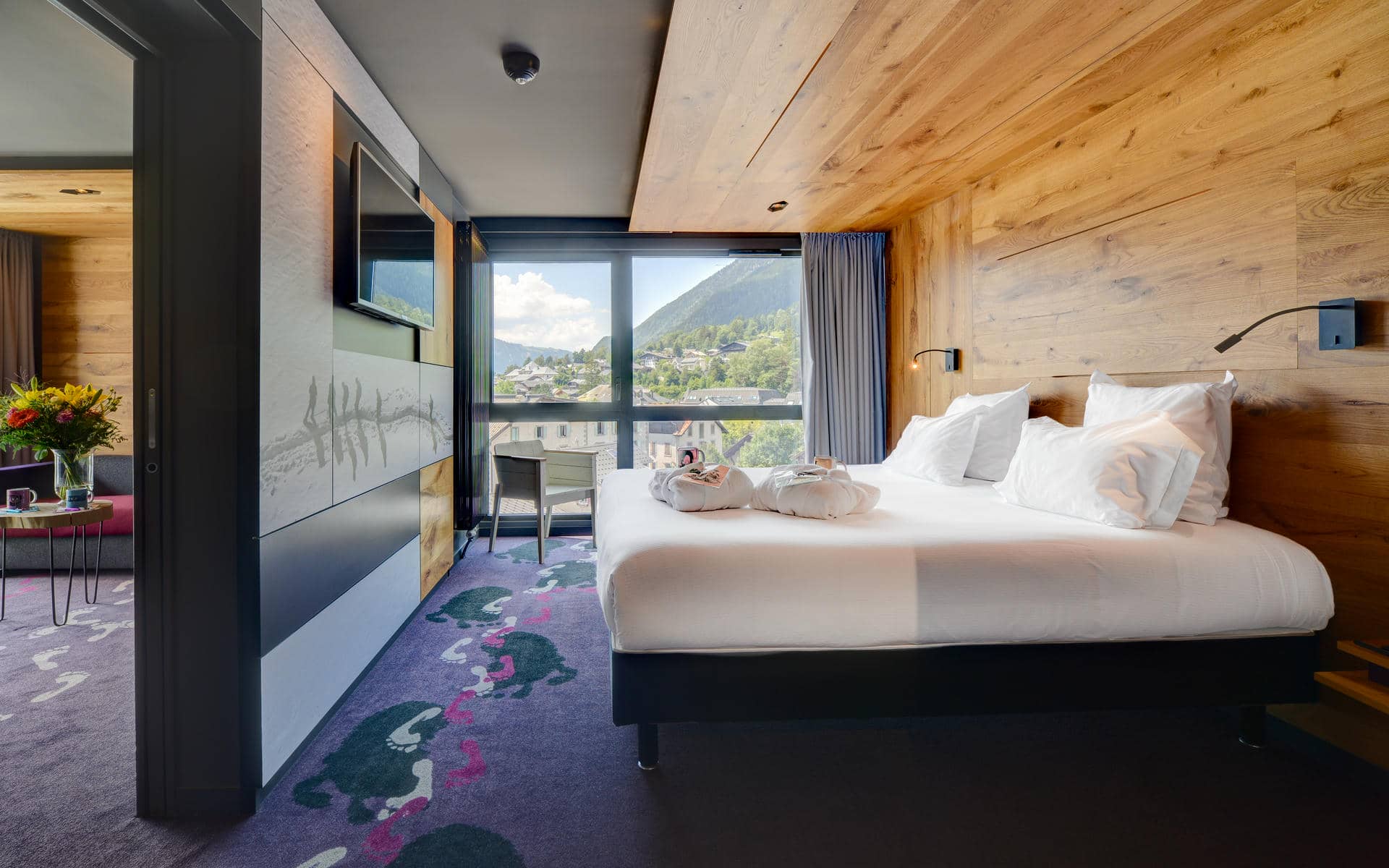 Chambre d'hôtel pour un séminaire à la montagne, avec un lit habillé de linge blanc et des serviettes pliées contre un mur en bois à droite, une moquette violette imprimée d'empreintes de pas vertes et roses au premier plan, une télévision murale et un panneau illustré de traces d'animaux à gauche, et un bureau avec une chaise près d'une grande fenêtre donnant sur les toits d'un village de montagne et des collines verdoyantes en arrière-plan.