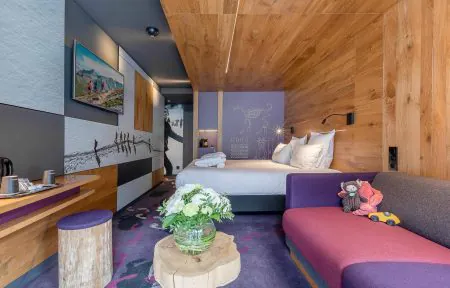 Chambre d'hôtel pour un séminaire à la montagne, avec un canapé banquette violet et gris parsemé de peluches à droite, un bureau en bois avec des tasses et une machine à café à gauche, une table d'appoint en tronc d'arbre portant un bouquet de fleurs blanches au premier plan, et un lit habillé de linge blanc contre un mur en bois avec une illustration de créature alpine « Dahu » sur un panneau mauve, sous une télévision murale en arrière-plan.
