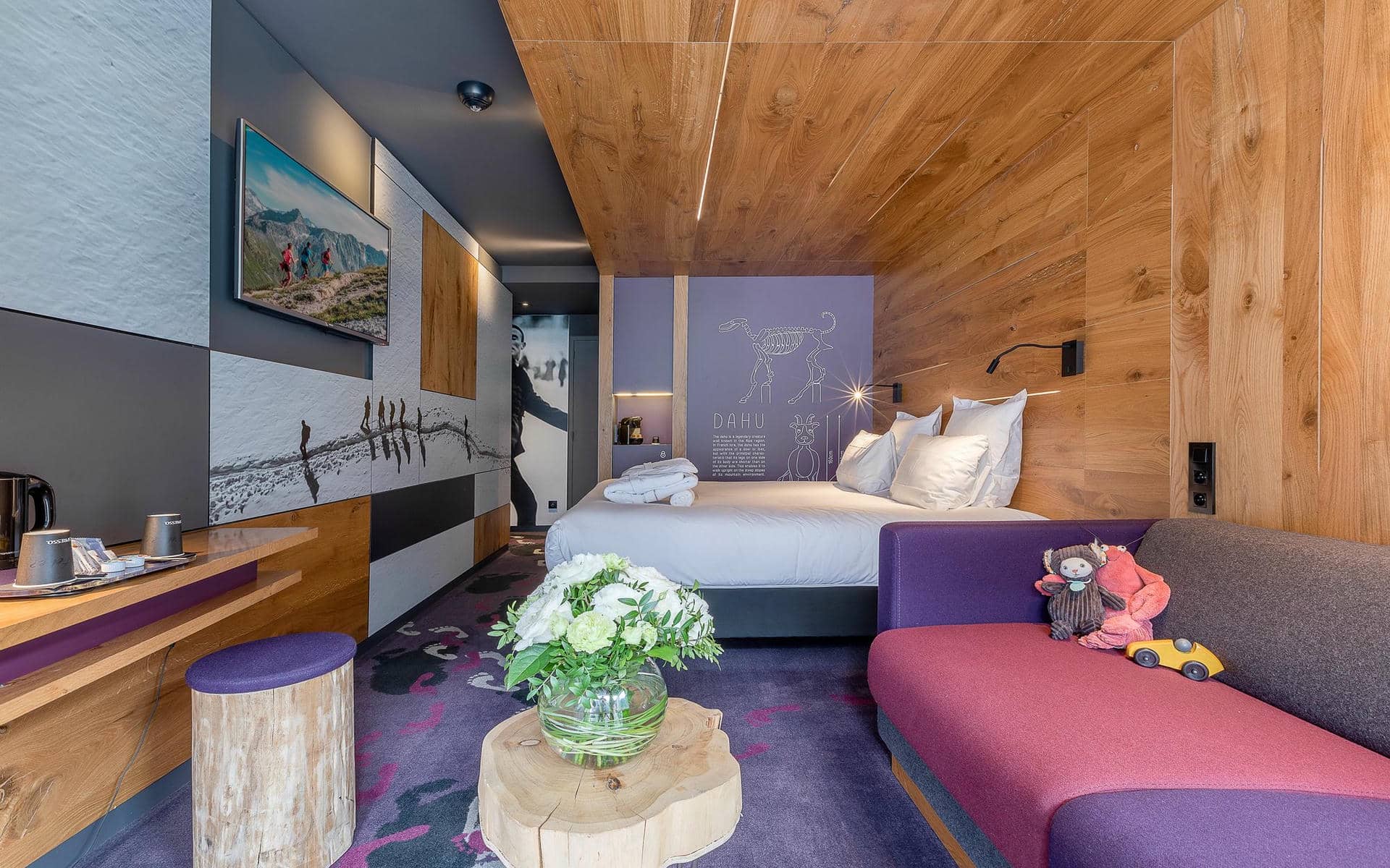 Chambre d'hôtel pour un séminaire à la montagne, avec un canapé banquette violet et gris parsemé de peluches à droite, un bureau en bois avec des tasses et une machine à café à gauche, une table d'appoint en tronc d'arbre portant un bouquet de fleurs blanches au premier plan, et un lit habillé de linge blanc contre un mur en bois avec une illustration de créature alpine « Dahu » sur un panneau mauve, sous une télévision murale en arrière-plan.