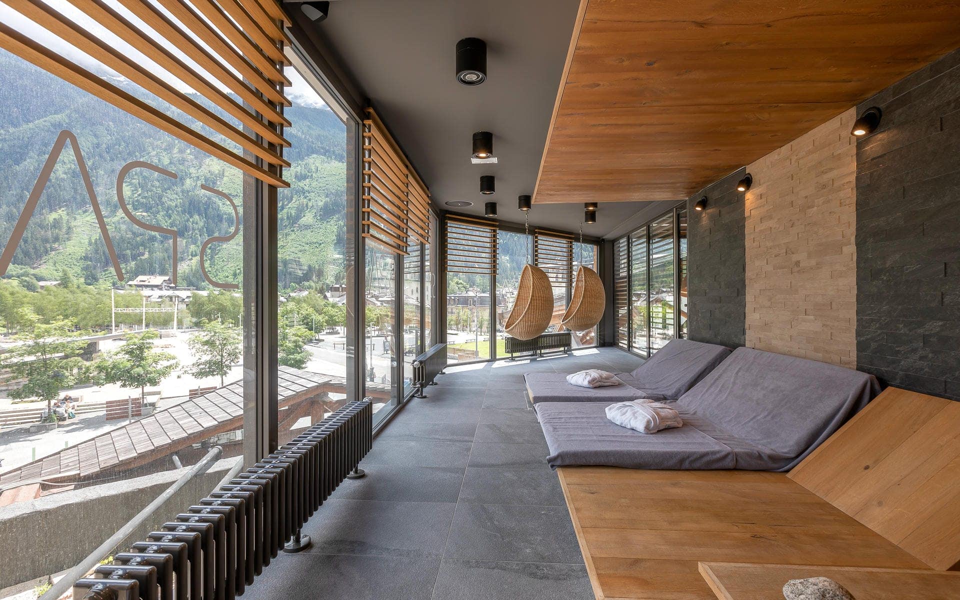 Espace détente du spa d'un hôtel avec transats matelassés gris et serviettes blanches pliées à droite, deux fauteuils suspendus en osier près de baies vitrées du sol au plafond à stores en lattes de bois donnant sur une vallée montagneuse au milieu, mur habillé de pierre à droite, et logo « Spa » gravé sur la vitre au premier plan à gauche, un espace de détente pour un séminaire bien-être.