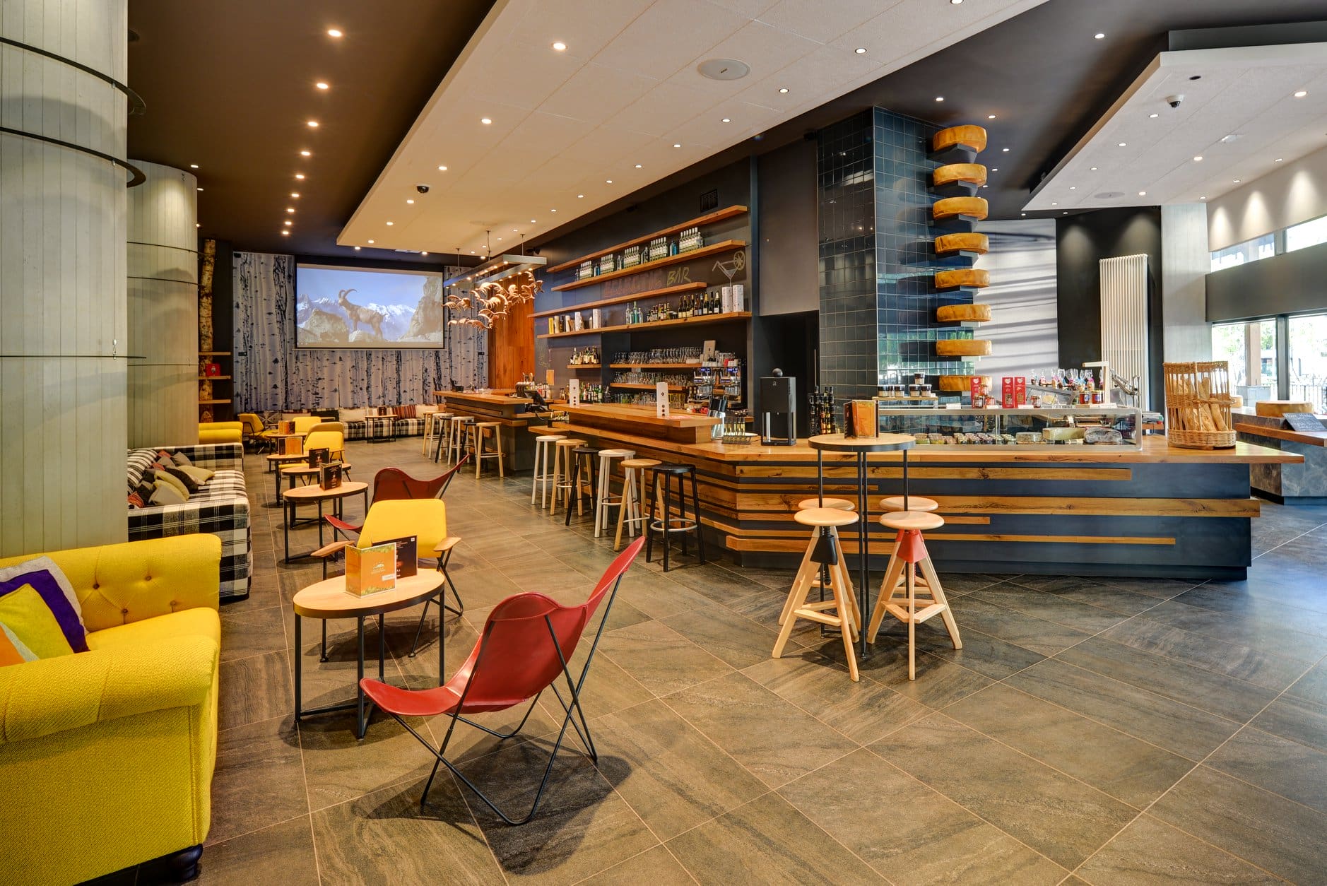 Bar-salon d'hôtel avec comptoir en bois et bleu foncé garni de bouteilles et verrerie au milieu, fauteuils papillon rouges et petites tables rondes au premier plan, canapé jaune et banquette à carreaux avec coussins à gauche, décor de meules de fromage empilées au-dessus d'une vitrine et tabourets hauts à droite, et écran de projection montrant une scène de bouquetin en montagne en arrière-plan, un espace bar et salon adapté à un séminaire à la montagne.
