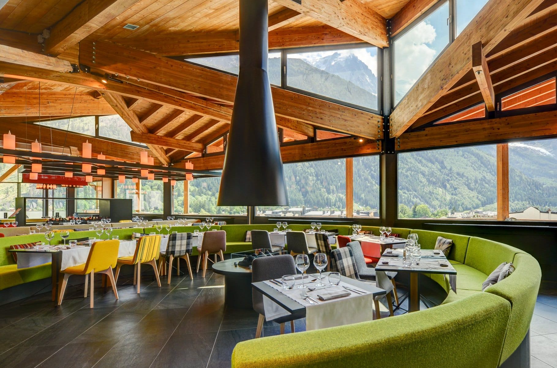Restaurant d'hôtel avec banquette verte incurvée entourant des tables nappées de blanc dressées de verres à vin et chaises à motifs colorés au premier plan, grande cheminée suspendue conique noire et suspensions en forme de lanternes au-dessus, et baies vitrées du sol au plafond dévoilant des montagnes boisées et un sommet enneigé en arrière-plan, un restaurant proposant une restauration sur place dans un cadre de séminaire à la montagne.