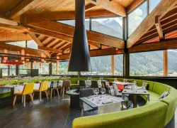Restaurant d'hôtel avec banquette verte incurvée entourant des tables nappées de blanc dressées de verres à vin et chaises à motifs colorés au premier plan, grande cheminée suspendue conique noire et suspensions en forme de lanternes au-dessus, et baies vitrées du sol au plafond dévoilant des montagnes boisées et un sommet enneigé en arrière-plan, un restaurant proposant une restauration sur place dans un cadre de séminaire à la montagne.