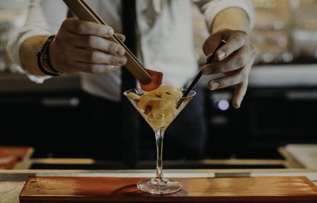 Afterwork gros plan sur les mains d'un barman utilisant des pinces pour garnir un verre à martini rempli de fruits et d'une tranche de fraise au centre, le verre posé sur une planche en bois foncé sur un comptoir de bar au premier plan, et un arrière-plan de bar tamisé avec bouteilles et étagères floues.