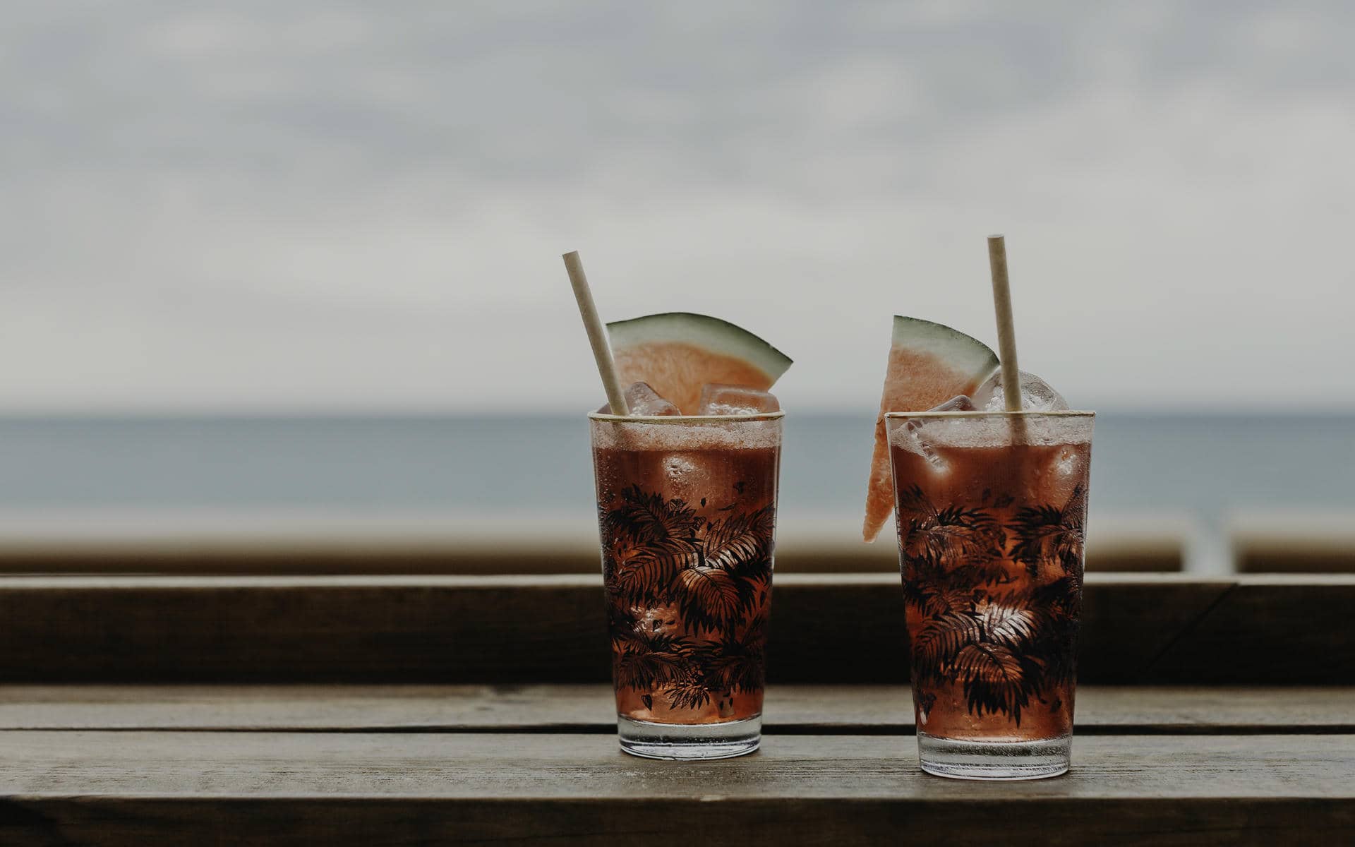Afterwork gros plan sur deux verres à motif de feuilles remplis d'une boisson rouge foncé, de glaçons, de tranches de pastèque et de pailles en papier sur un rebord en bois au premier plan, et un horizon marin flou sous un ciel gris en arrière-plan.
