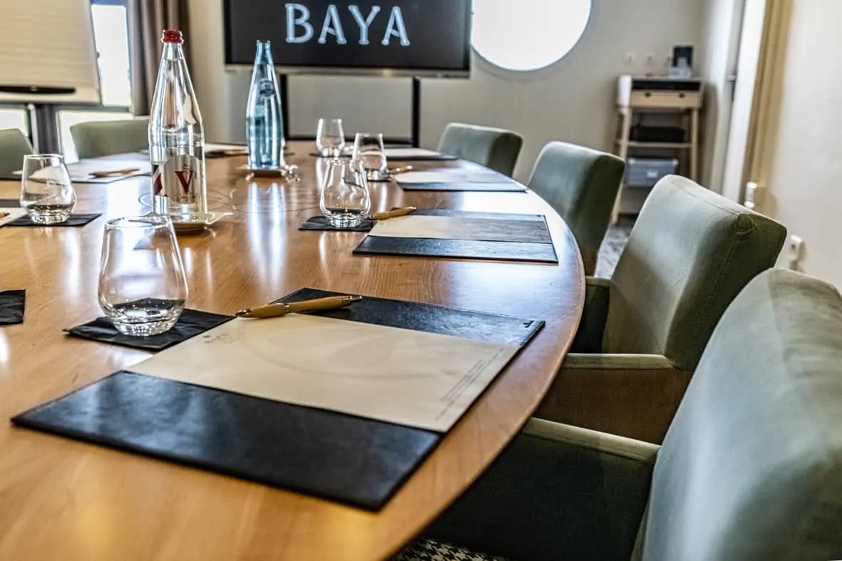 Gros plan sur une table de réunion ovale en bois dressée avec des sous-mains en cuir noir, des stylos, des verres et des bouteilles d'eau au premier plan, des fauteuils en cuir vert sauge disposés autour de la table à droite, et un écran numérique noir affichant un logo sur le mur en arrière-plan, adapté à une location de salle.
