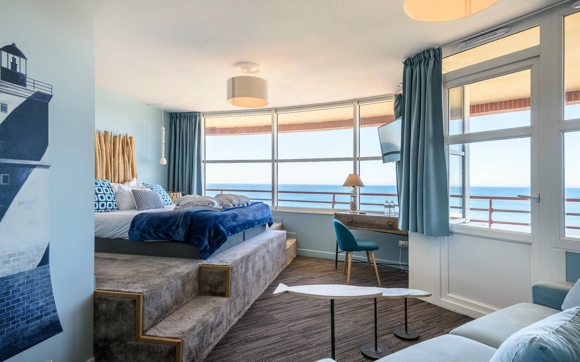 Chambre d'hôtel avec estrade de lit surélevée à tête de lit façon bois flotté et linge de lit bleu et blanc à gauche, fresque murale de phare à proximité, petites tables basses en forme de poisson et canapé bleu au premier plan, bureau en bois avec chaise bleu canard et télévision murale au milieu, et baies vitrées du sol au plafond et porte-fenêtre donnant sur la mer en arrière-plan, une chambre adaptée à un séminaire en bord de mer.